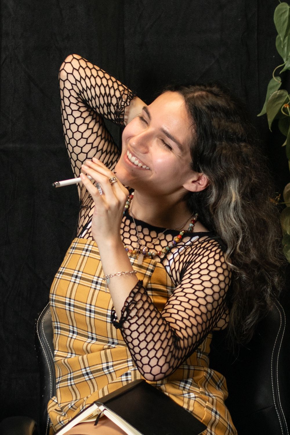 Happy person wearing yellow plaid vest and fishnet sleeves poses with long dark hair against black background.