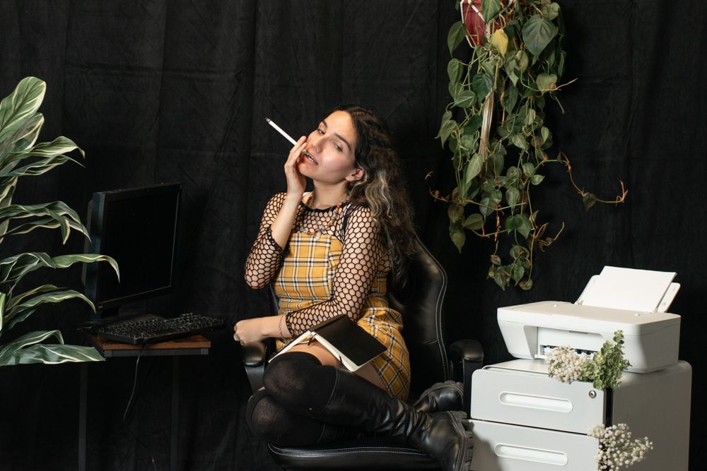 Person in vintage yellow plaid outfit sits contemplatively with cigarette among decorative boxes and plants.