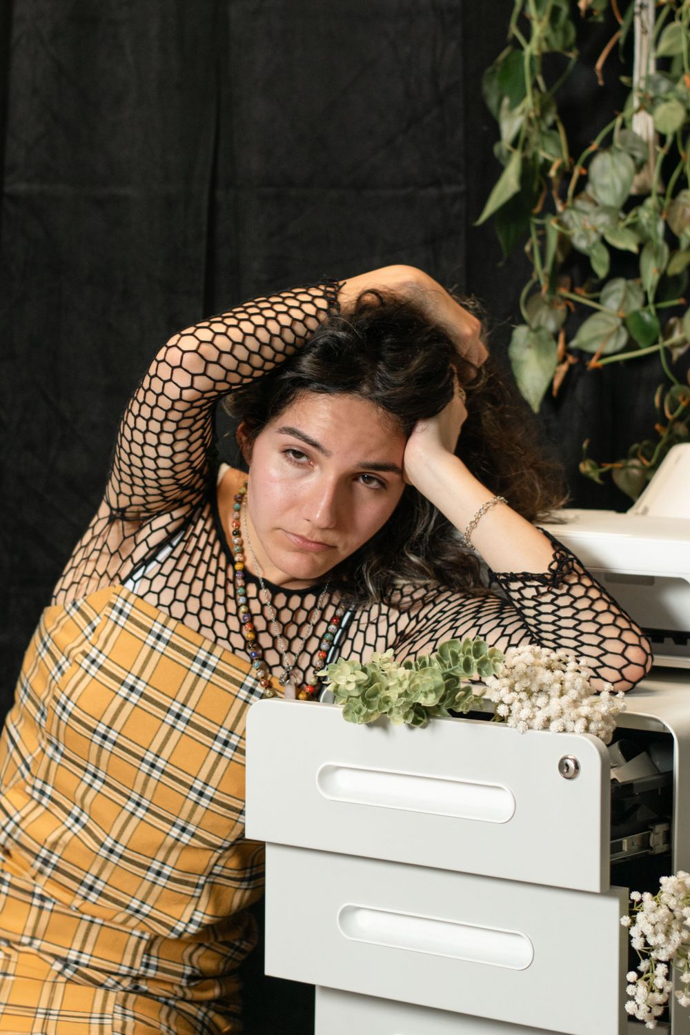 Person in geometric patterned top and plaid vest leans on white filing cabinet with dried flowers.