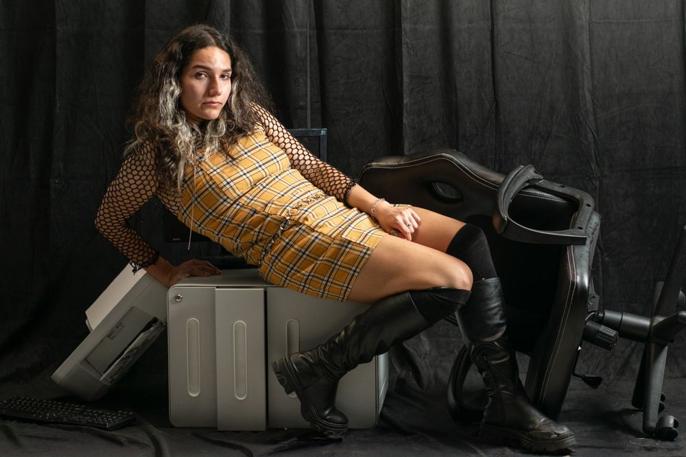 A person in a yellow plaid dress poses on silver luggage cases next to tall black boots against a dark background.