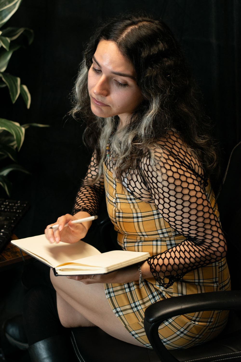 A person in a plaid dress and fishnet sleeves sits reading a book in moody lighting with plants in the background.