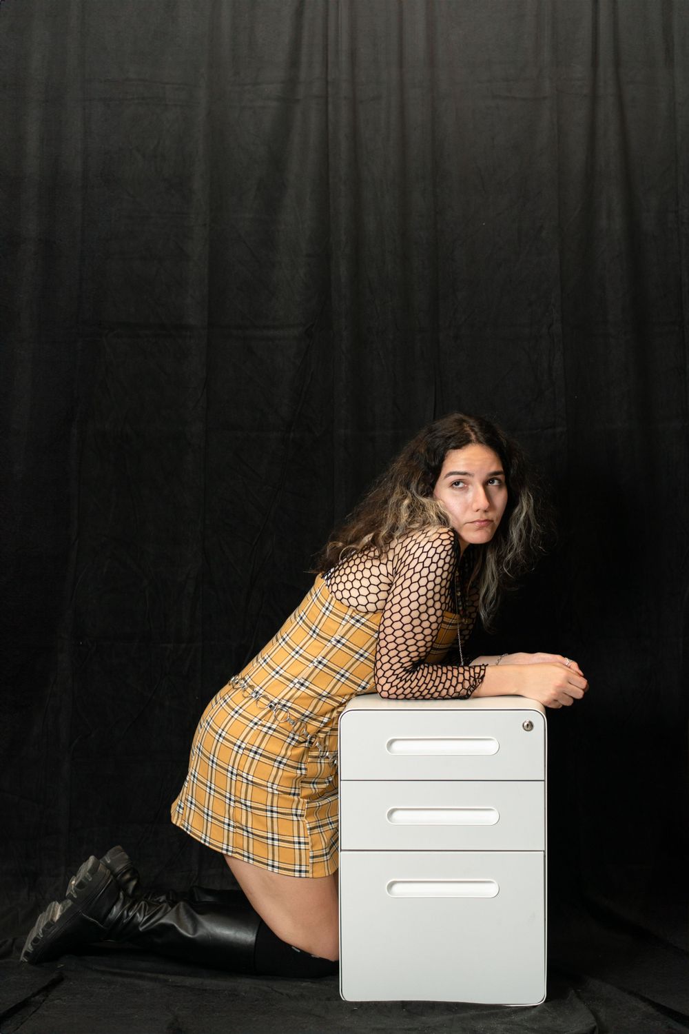Fashion portrait with plaid dress and mesh top posed against black backdrop beside white filing cabinet.