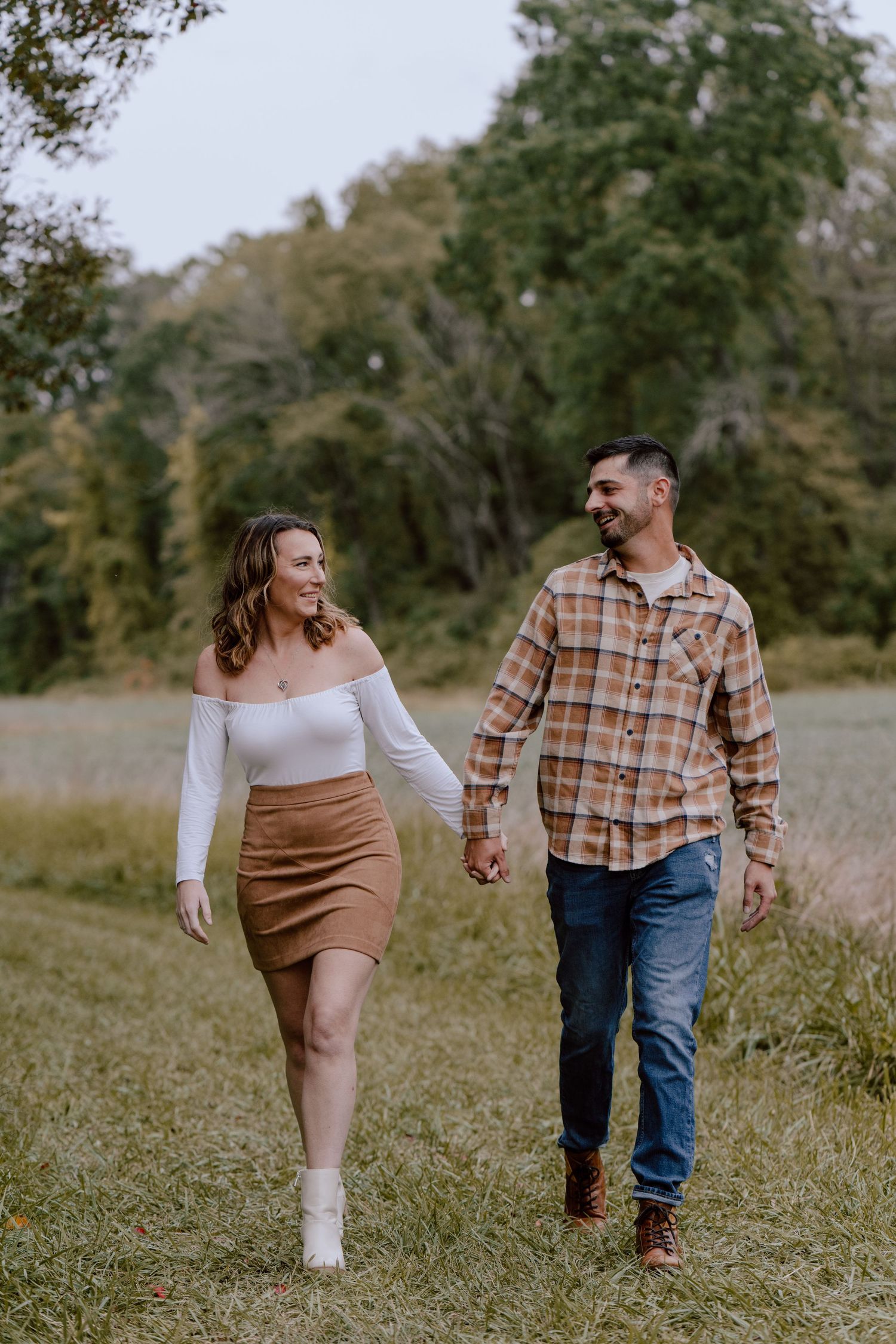 A couple walks hand in hand through a grassy field on an autumn day.