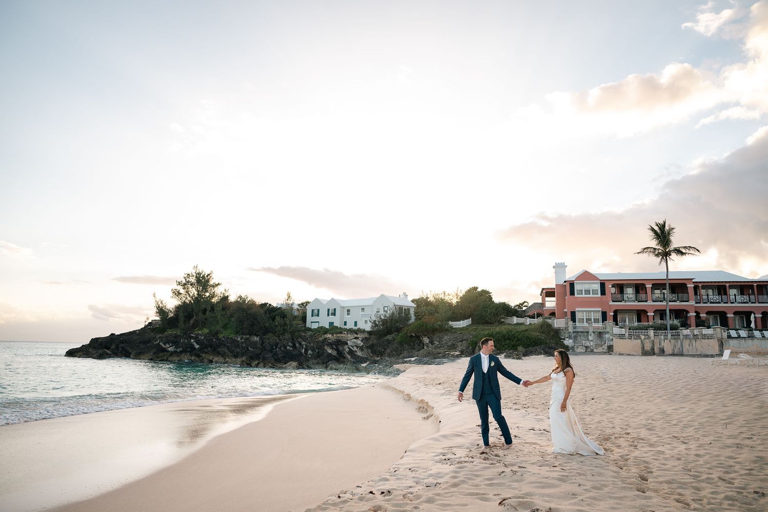 A couple in wedding attire walks along a scenic beach at sunset with palm trees and resort buildings in the background.