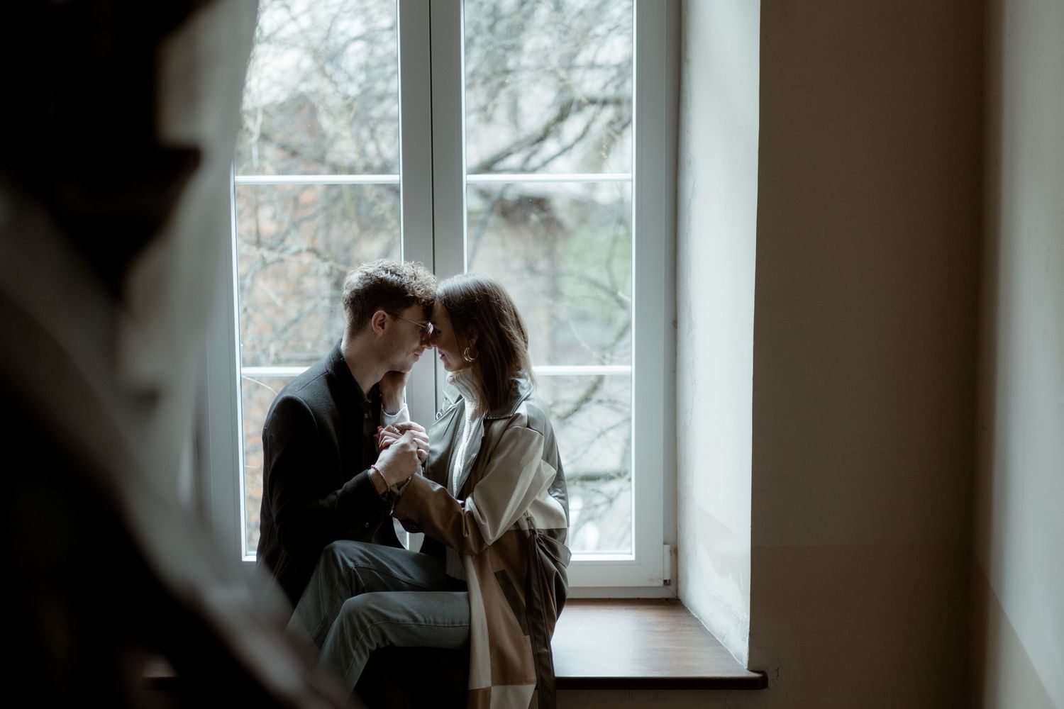 Two people share an intimate moment while sitting by a bright window, bathed in soft natural light.