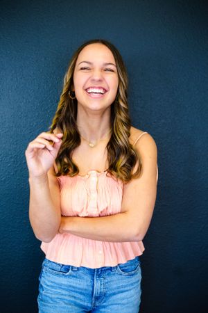 A joyful expression radiates warmth as a senior girl in a peach-colored top and jeans stands against a dark blue wall in a senior photo