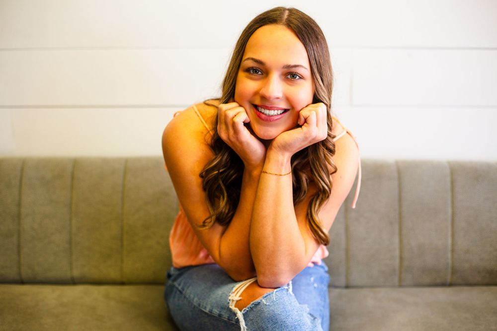 A cheerful senior wearing jeans and a peach-colored top sits casually on a couch with hands under their chin in Central City photo studio.