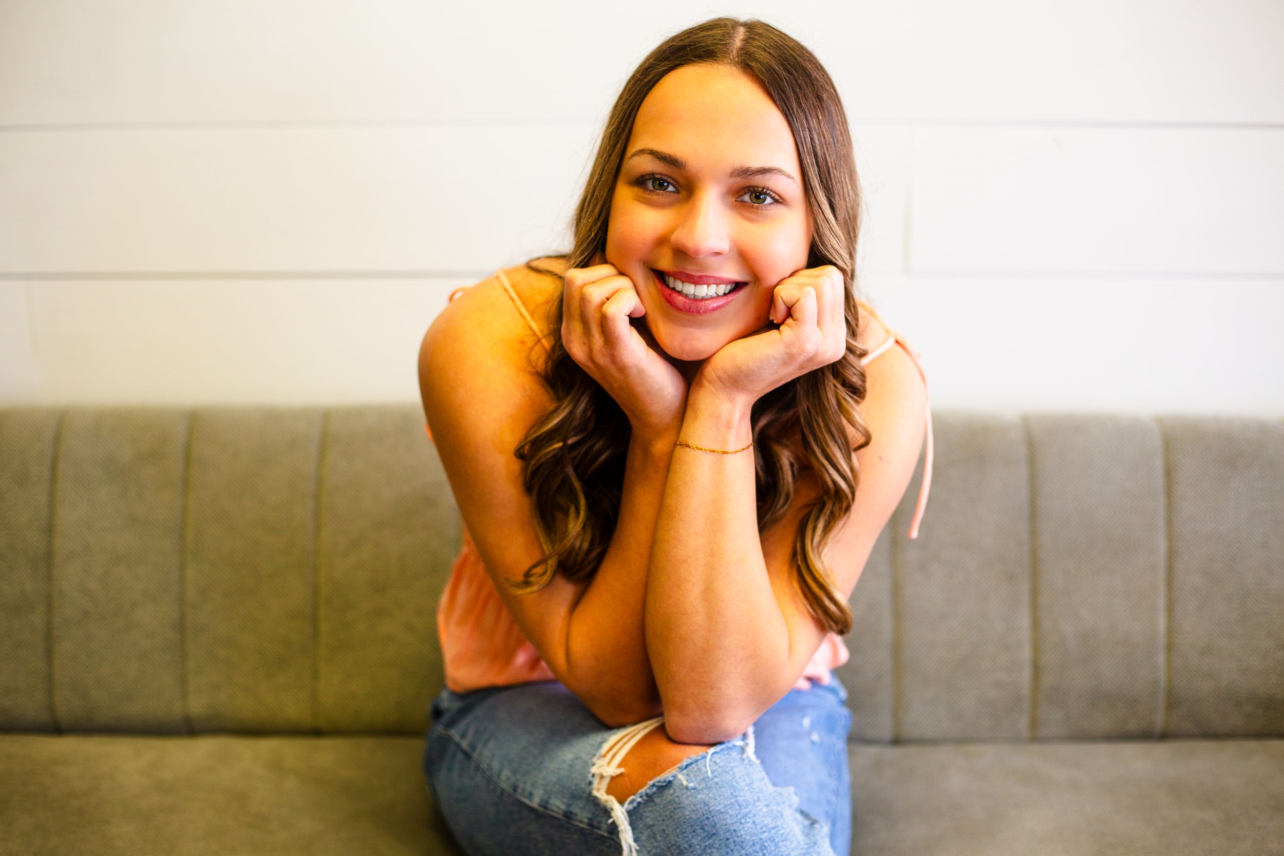 A cheerful senior wearing jeans and a peach-colored top sits casually on a couch with hands under their chin in Central City photo studio.