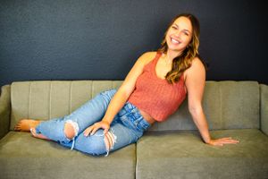 Senior girl in Central City photography studio lounges casually on a gray couch wearing a coral tank top, distressed jeans and tan heels.