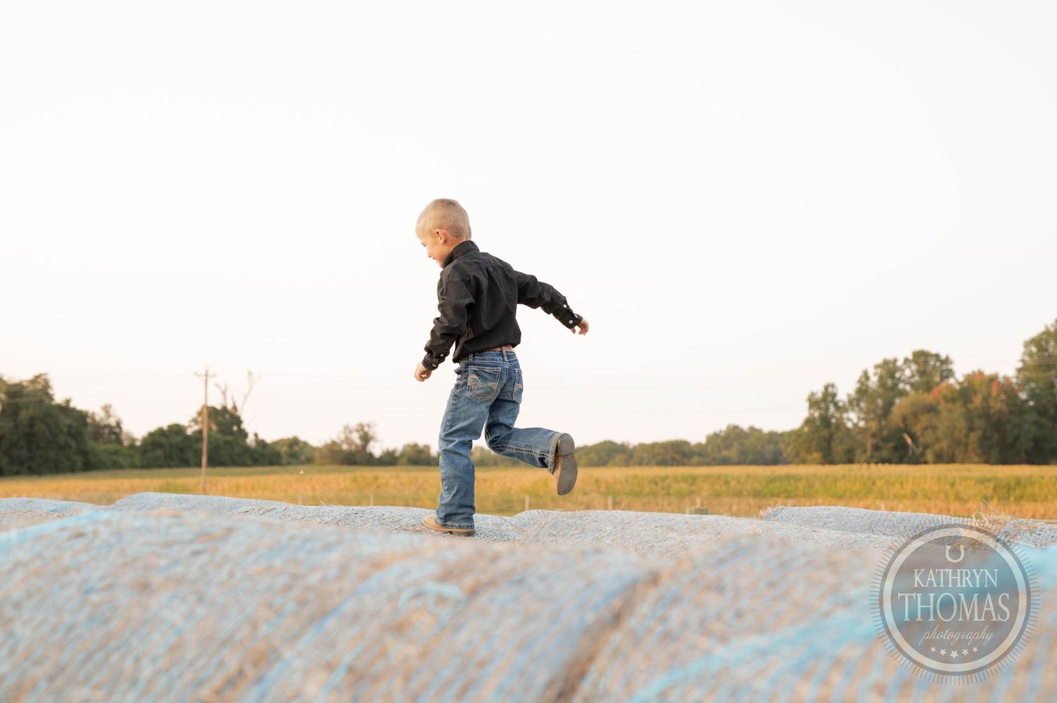 Child running playfully on large hay bales in a rural farm field at sunset.