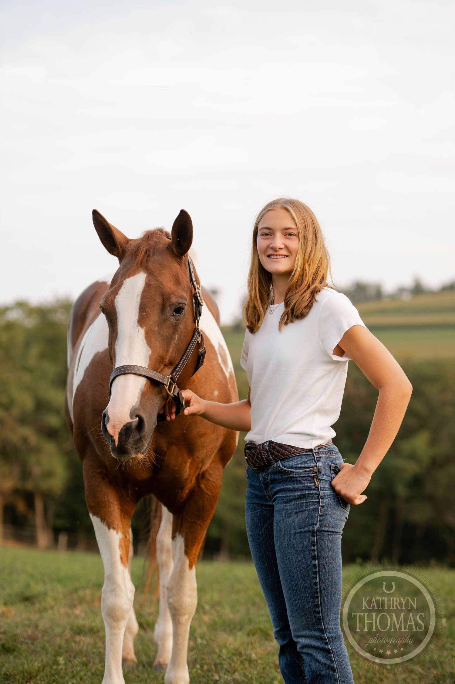 A paint horse with a white blaze stands in a sunny pasture with someone in a white t-shirt and jeans.