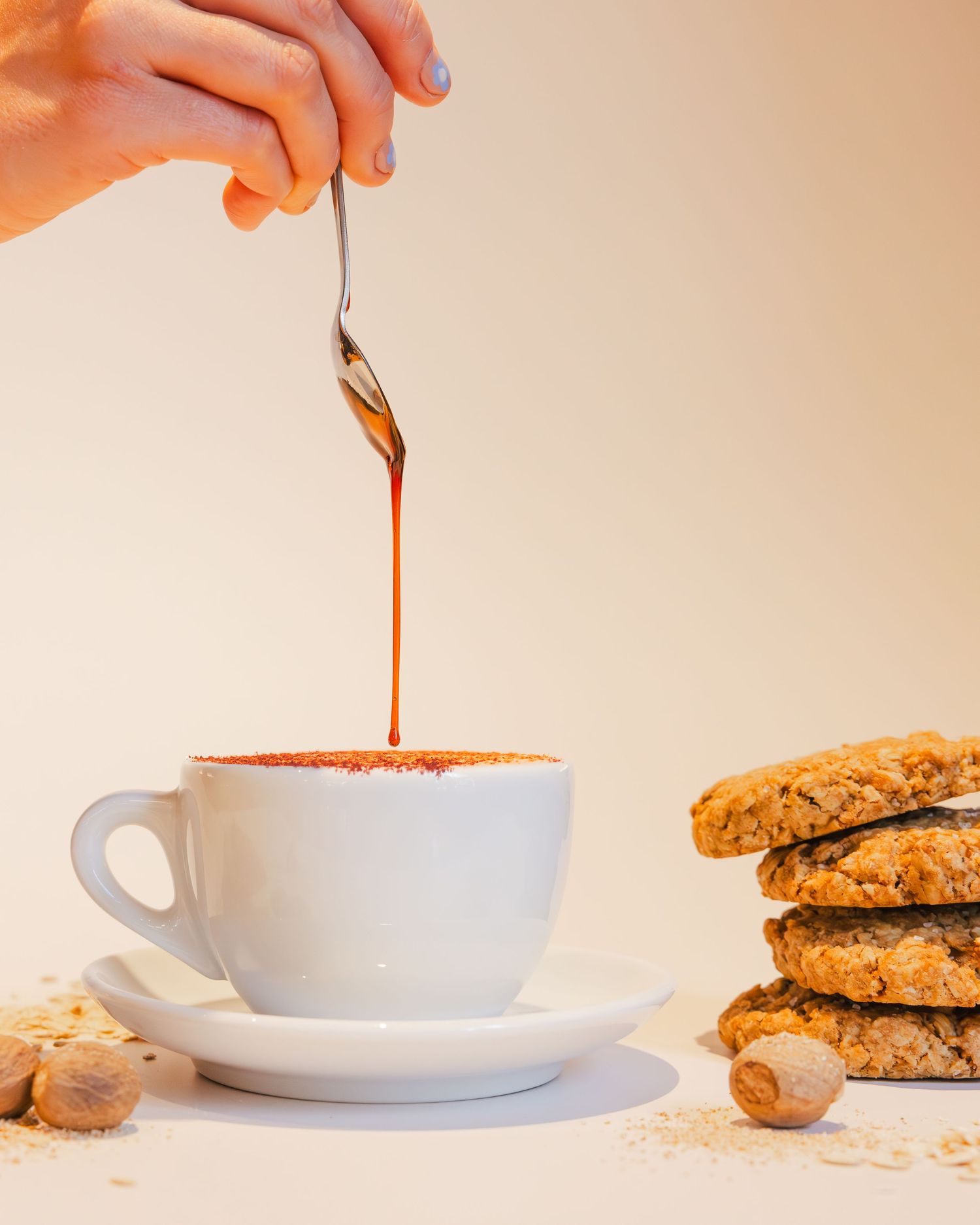 A hand pours syrup into a white coffee cup next to stacked chocolate chip cookies and scattered nuts on a light surface.