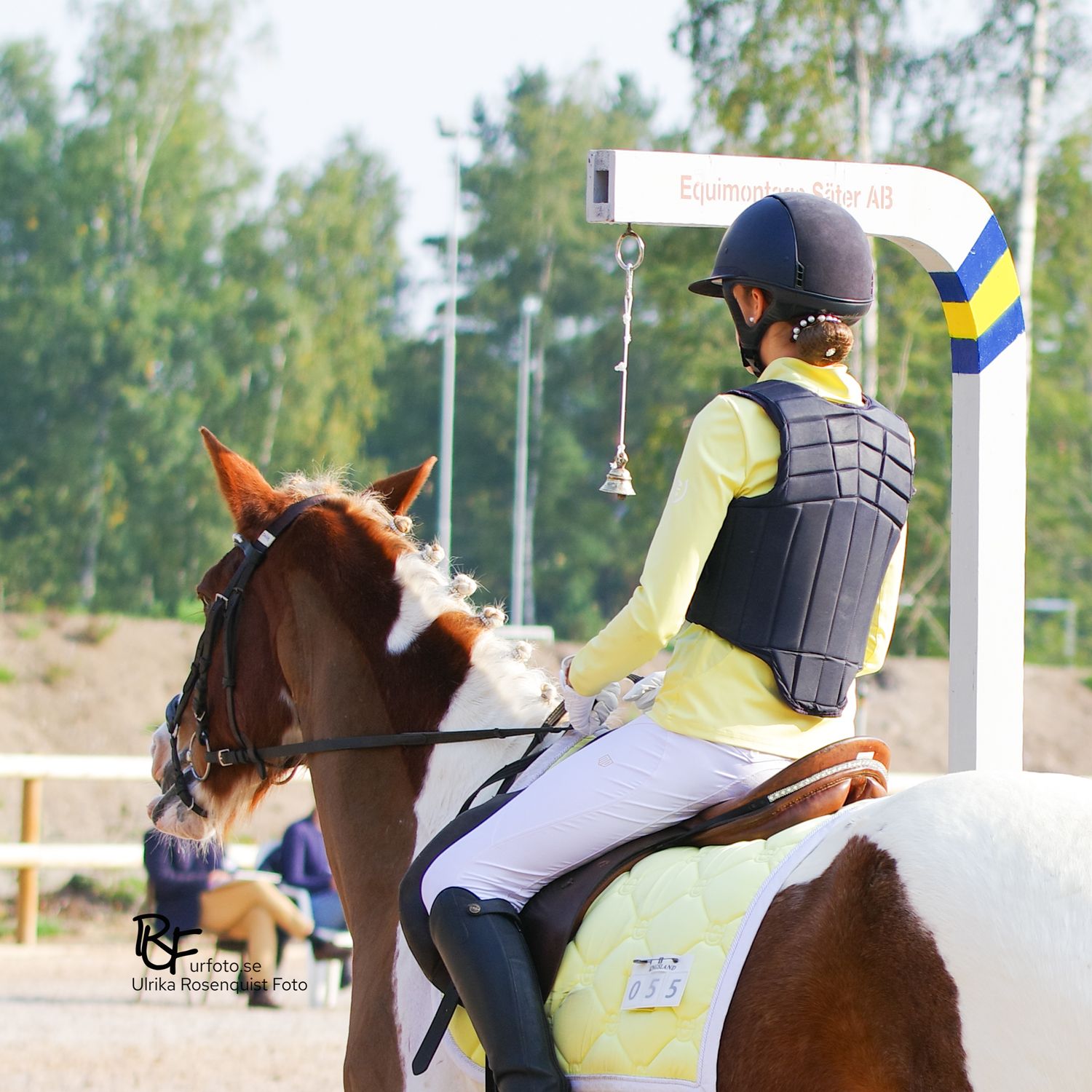 Ryttare i säkerhetsutrustning och gul topp på en brun och vit häst Working Equtation på utomhusarena.