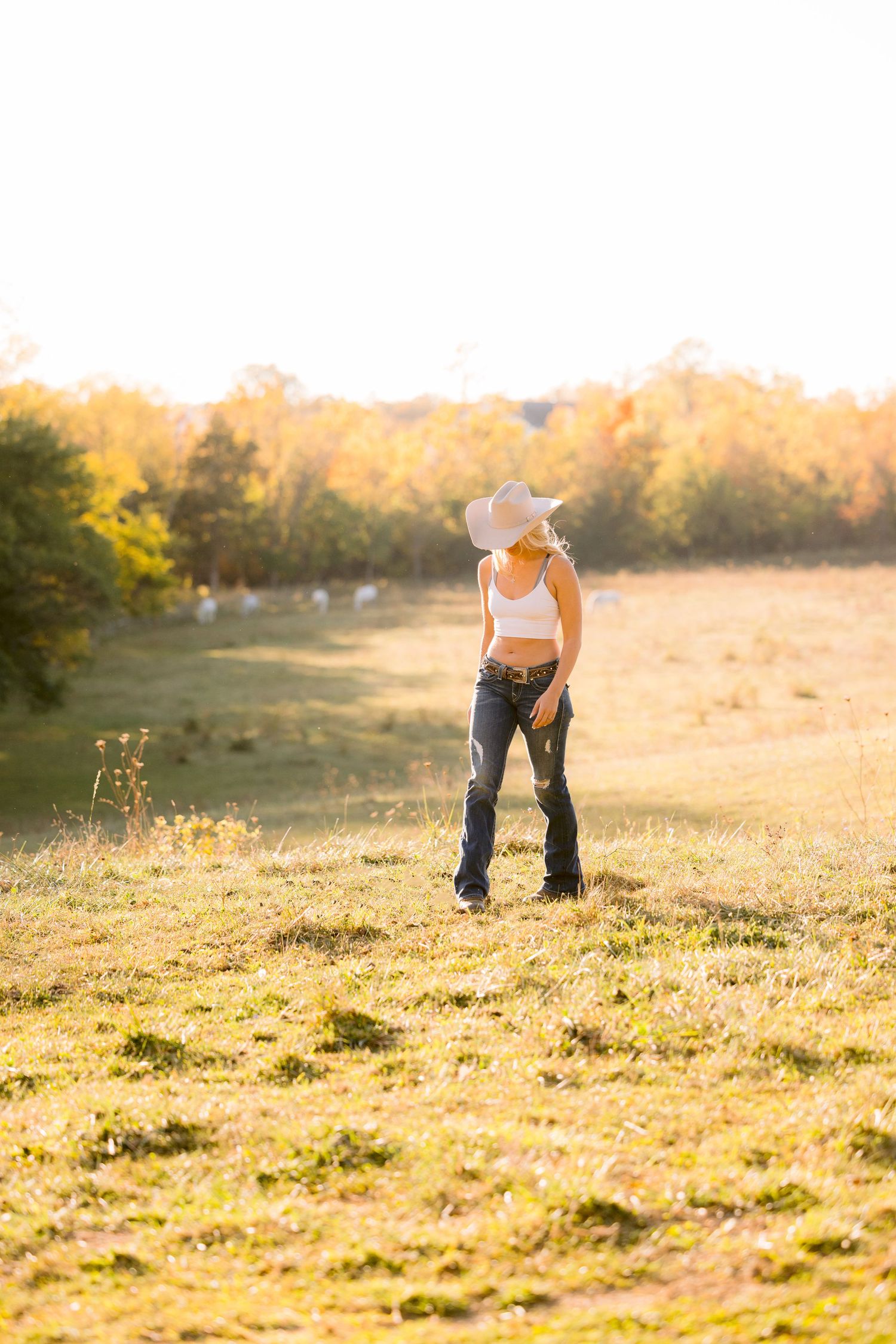 Person in white tank top and jeans stands in golden sunset field surrounded by autumn trees.
