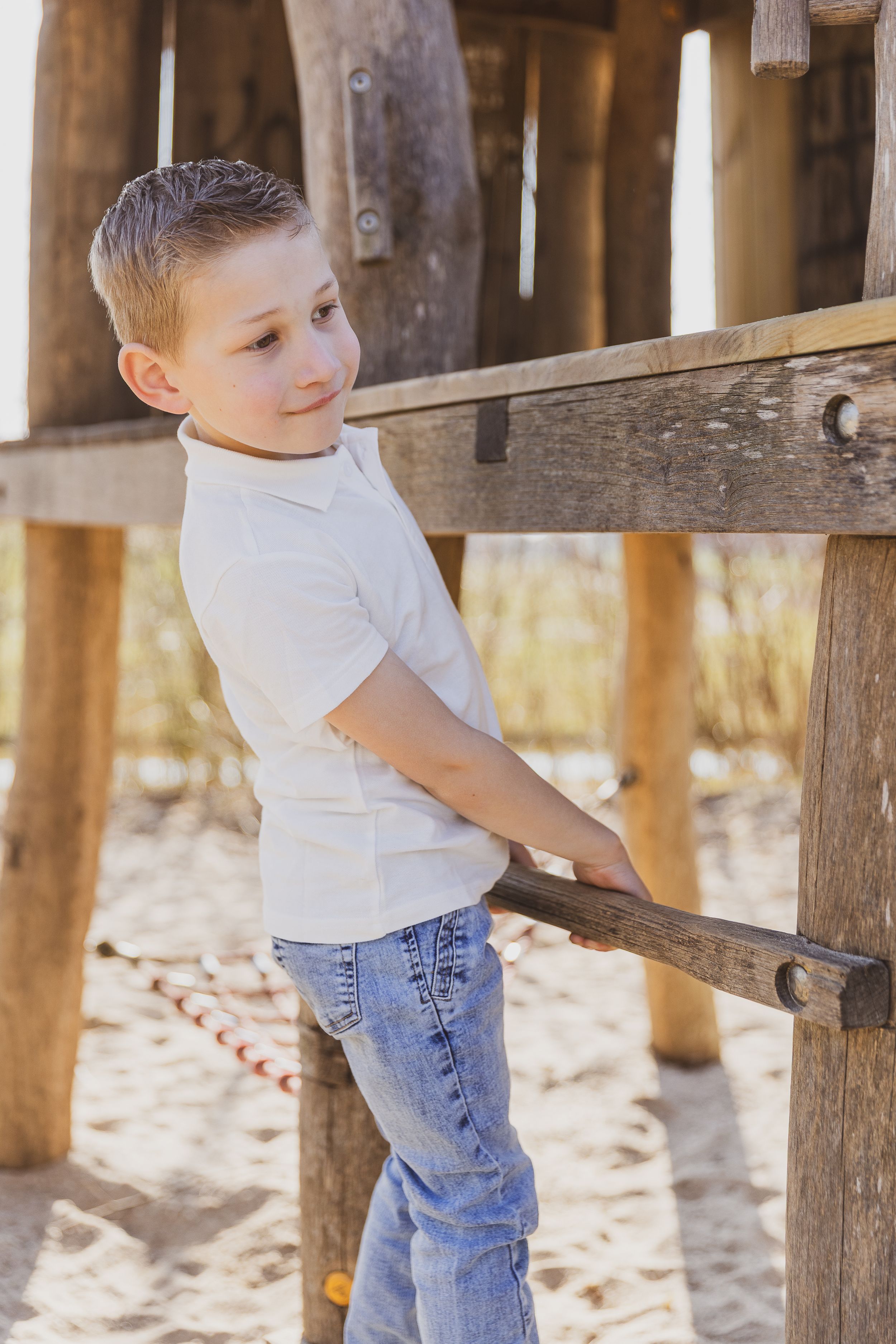 Young child in white shirt and jeans stands against wooden structure outdoors. gezinsshoot