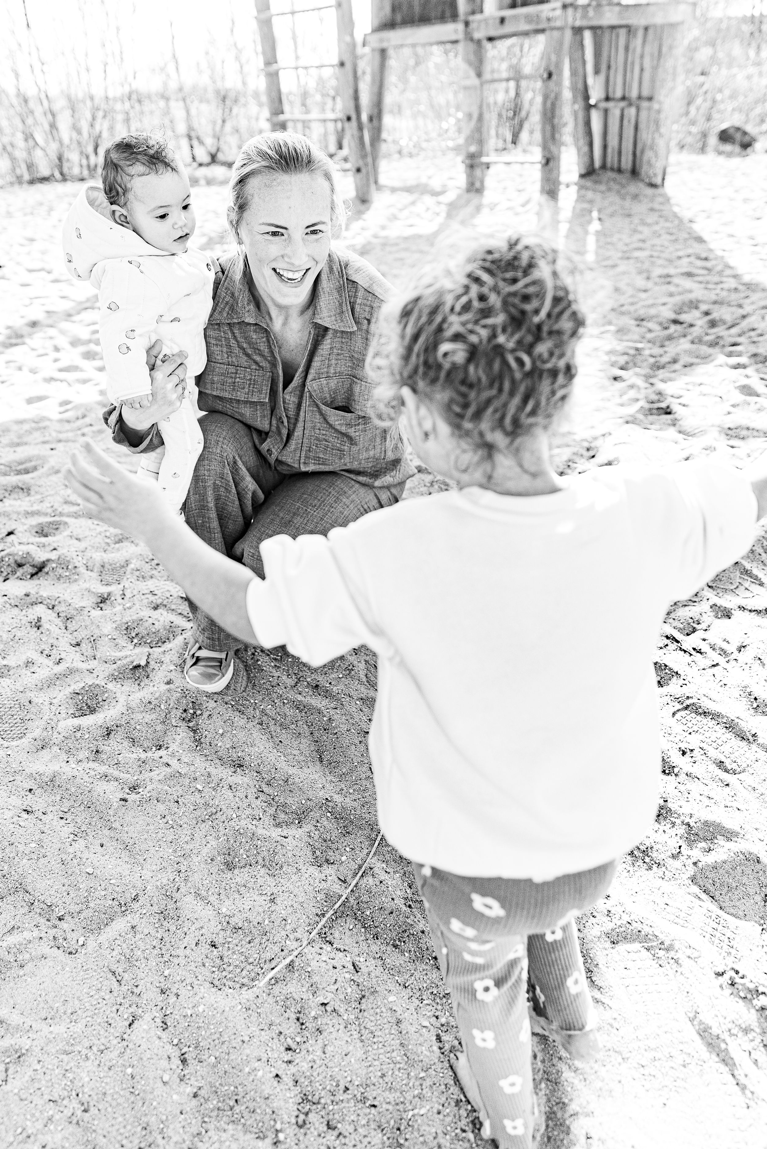 Black and white photo of family playing together in sandy outdoor area. Gezinsfotografie.