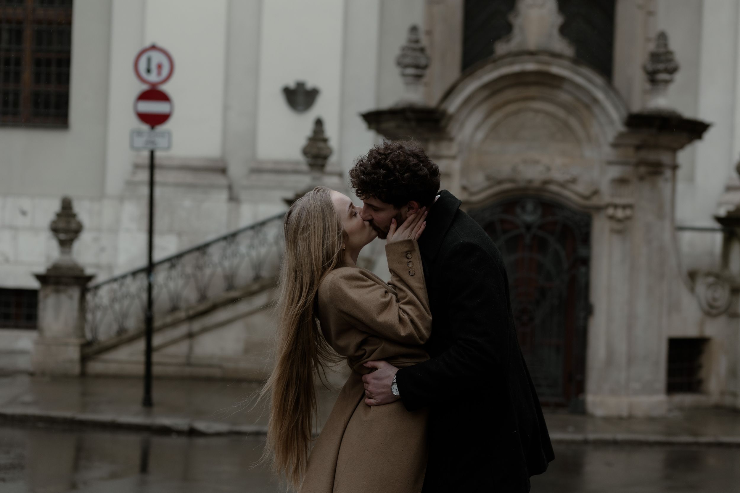 A romantic couple sharing a tender kiss outside an ornate historic building entrance with stone stairs.