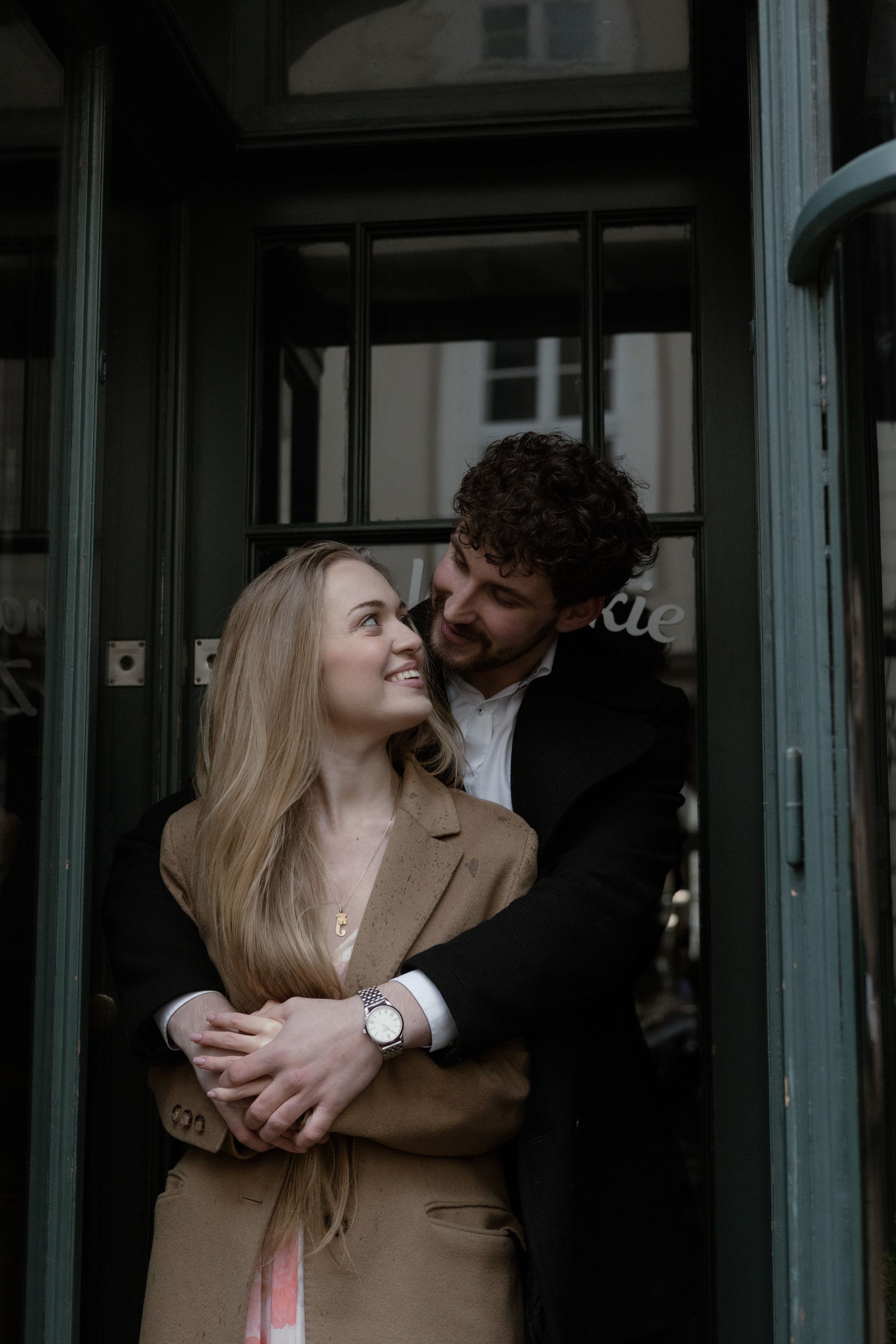 A couple shares an intimate moment in a doorway, with her wearing a tan coat and him in dark attire.