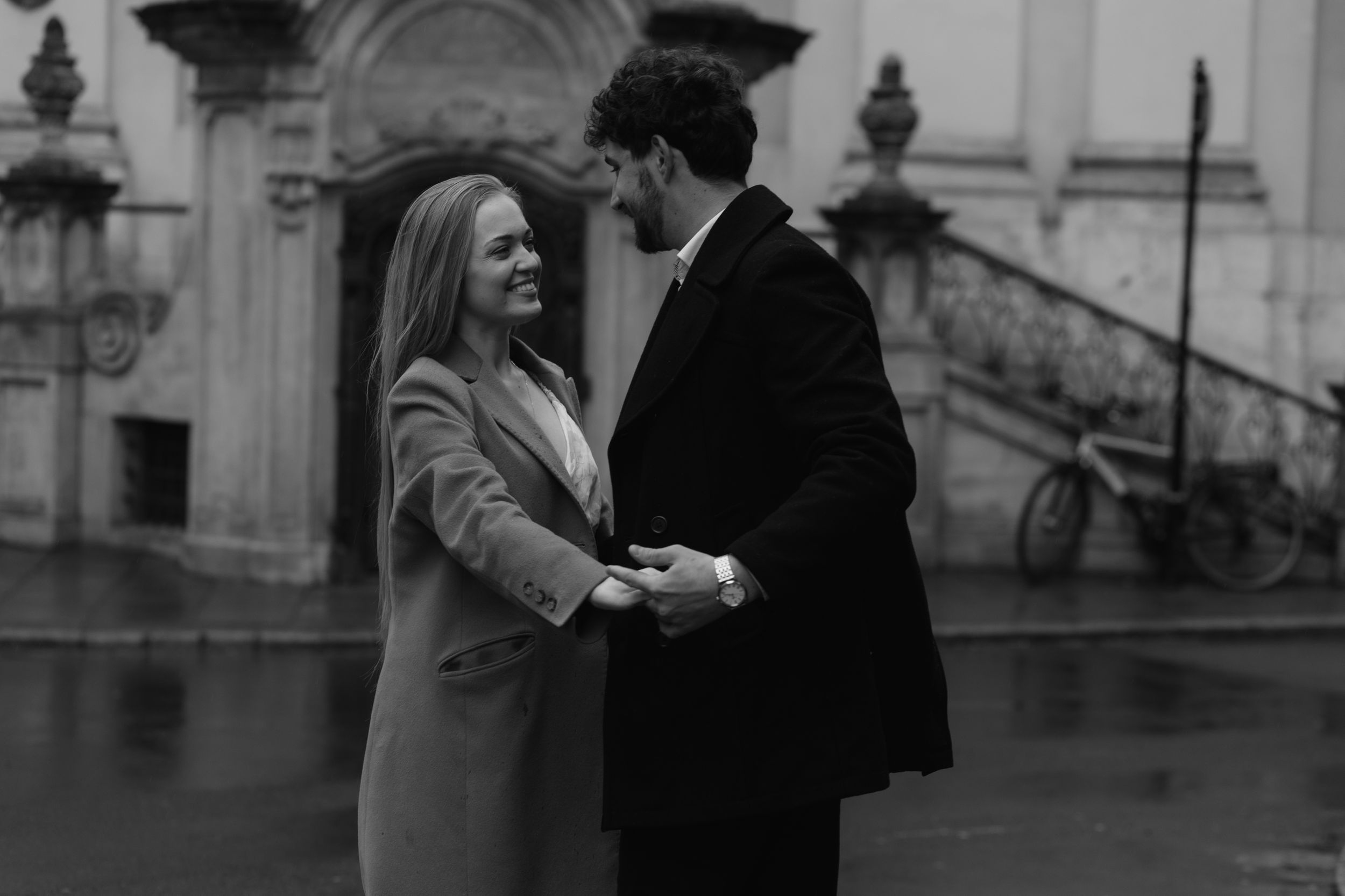 A romantic couple shares an intimate moment on a rainy day in front of historic architecture.