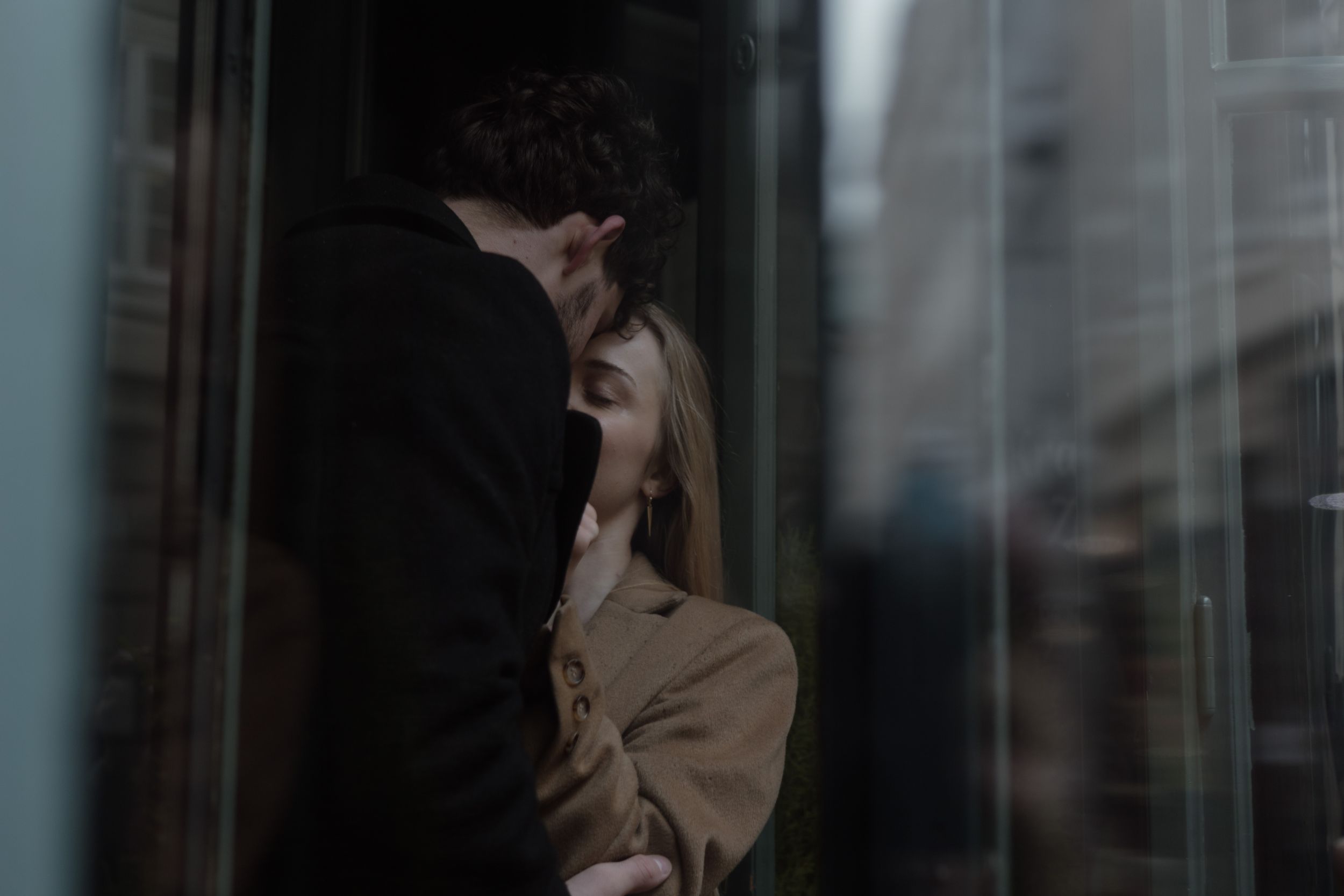 Someone in a beige coat stands by a window in dark moody lighting with their hand raised to their face.