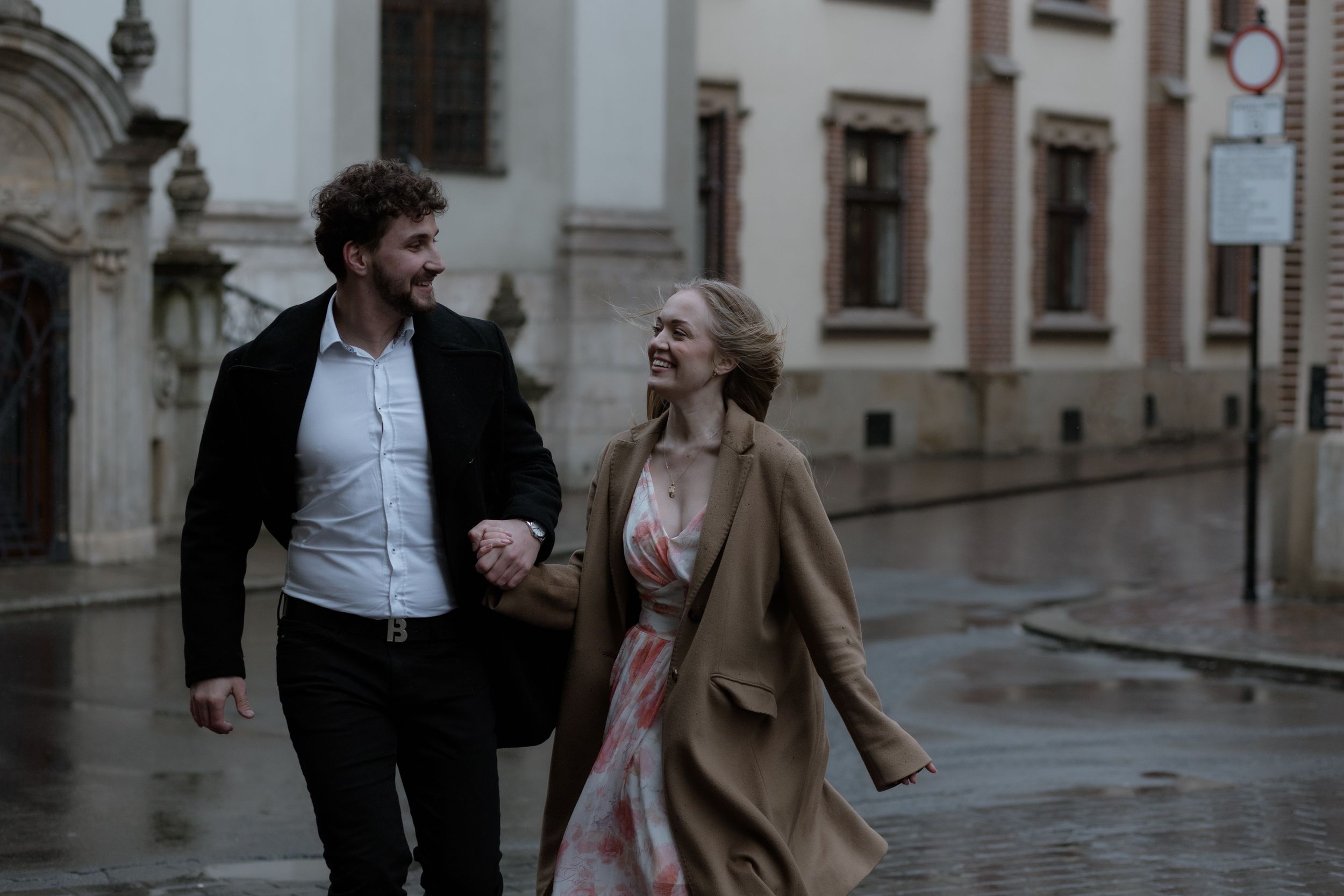 A couple walks together laughing on a rainy day in a historic European city street with classic architecture.