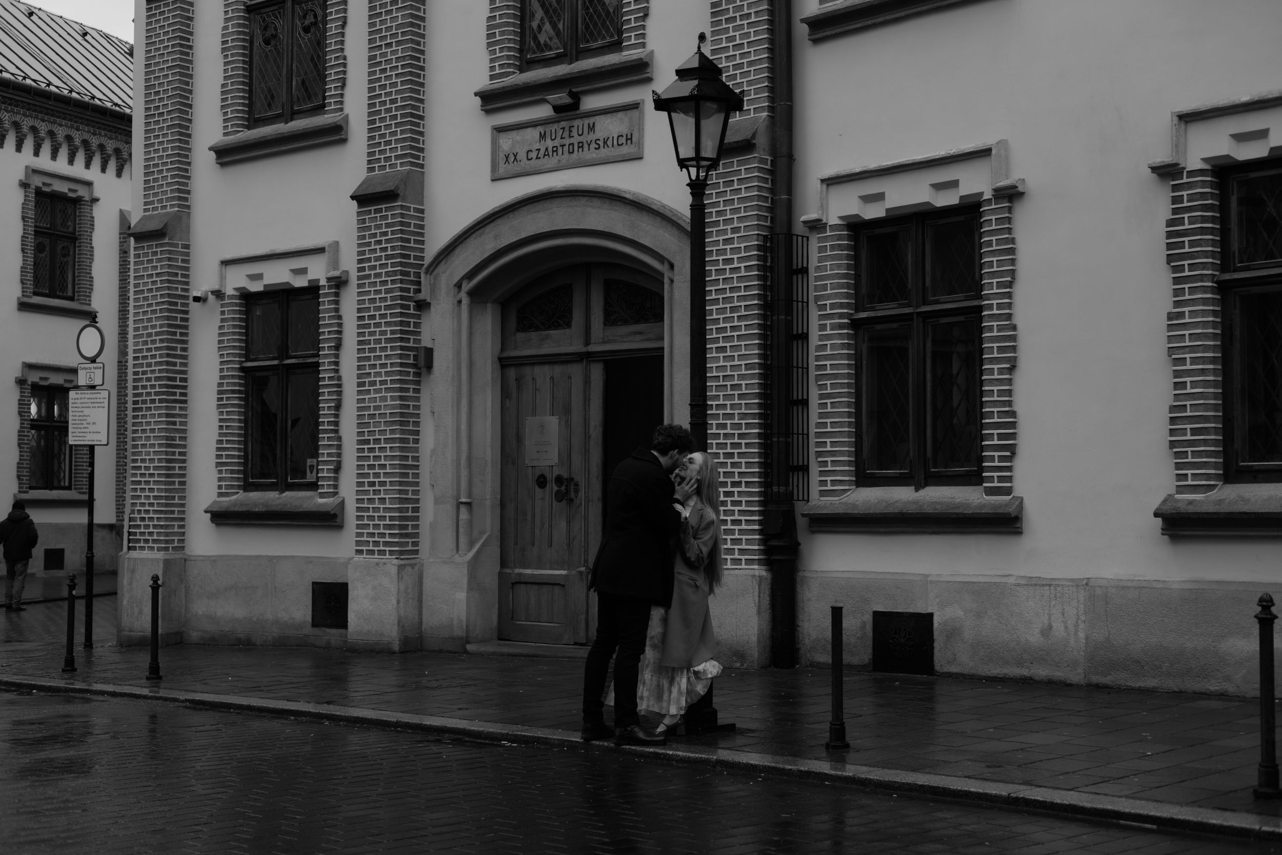 A black and white photo of a couple standing in the rain on a cobblestone street next to a vintage building.