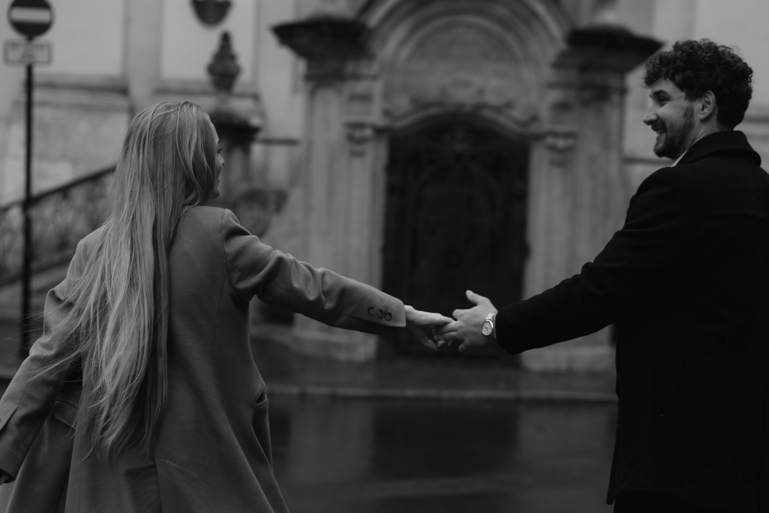 A couple holds hands at arms length in a romantic black and white moment outside a historic building.