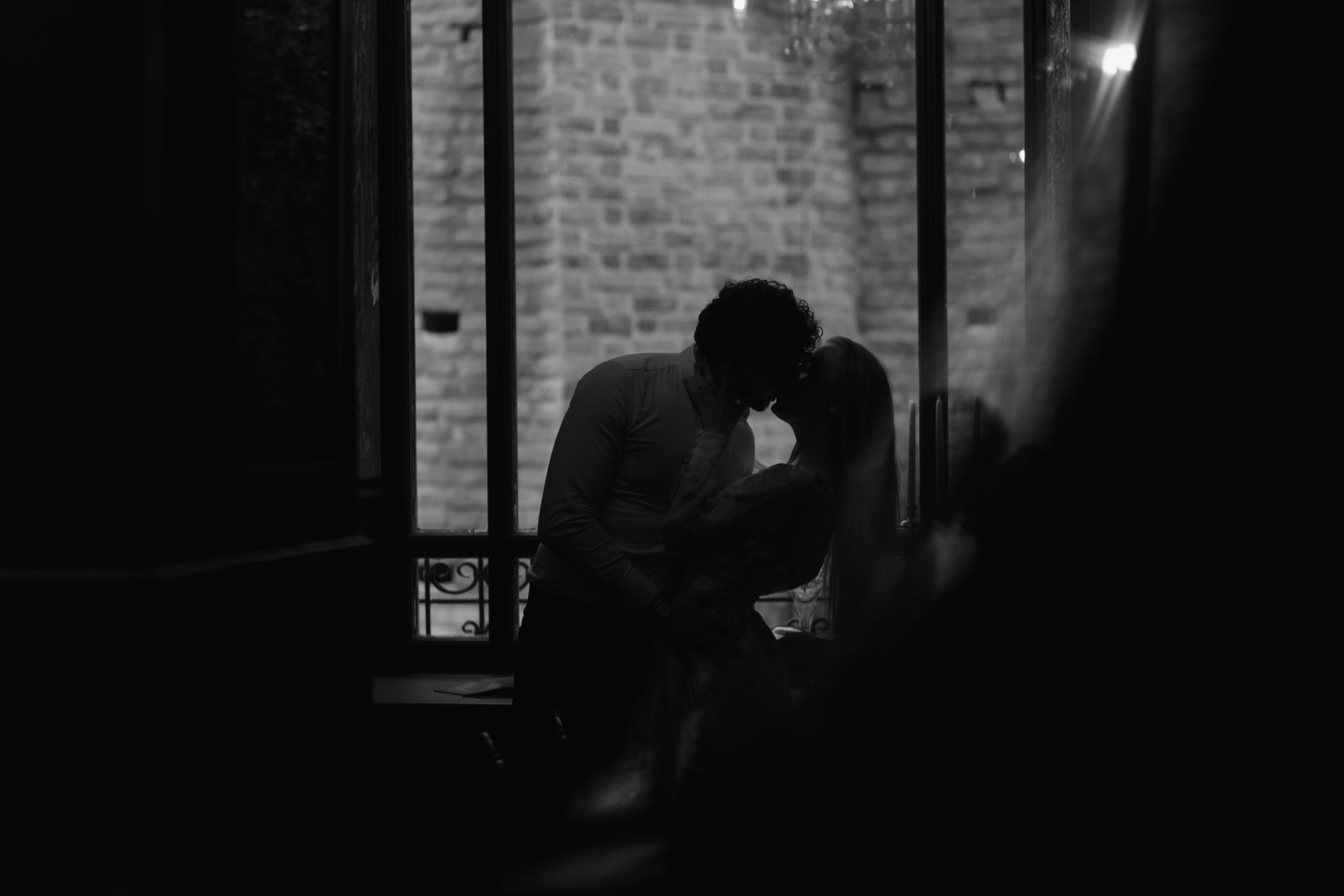 A silhouetted couple shares an intimate moment in a dimly lit room with brick walls in the background.