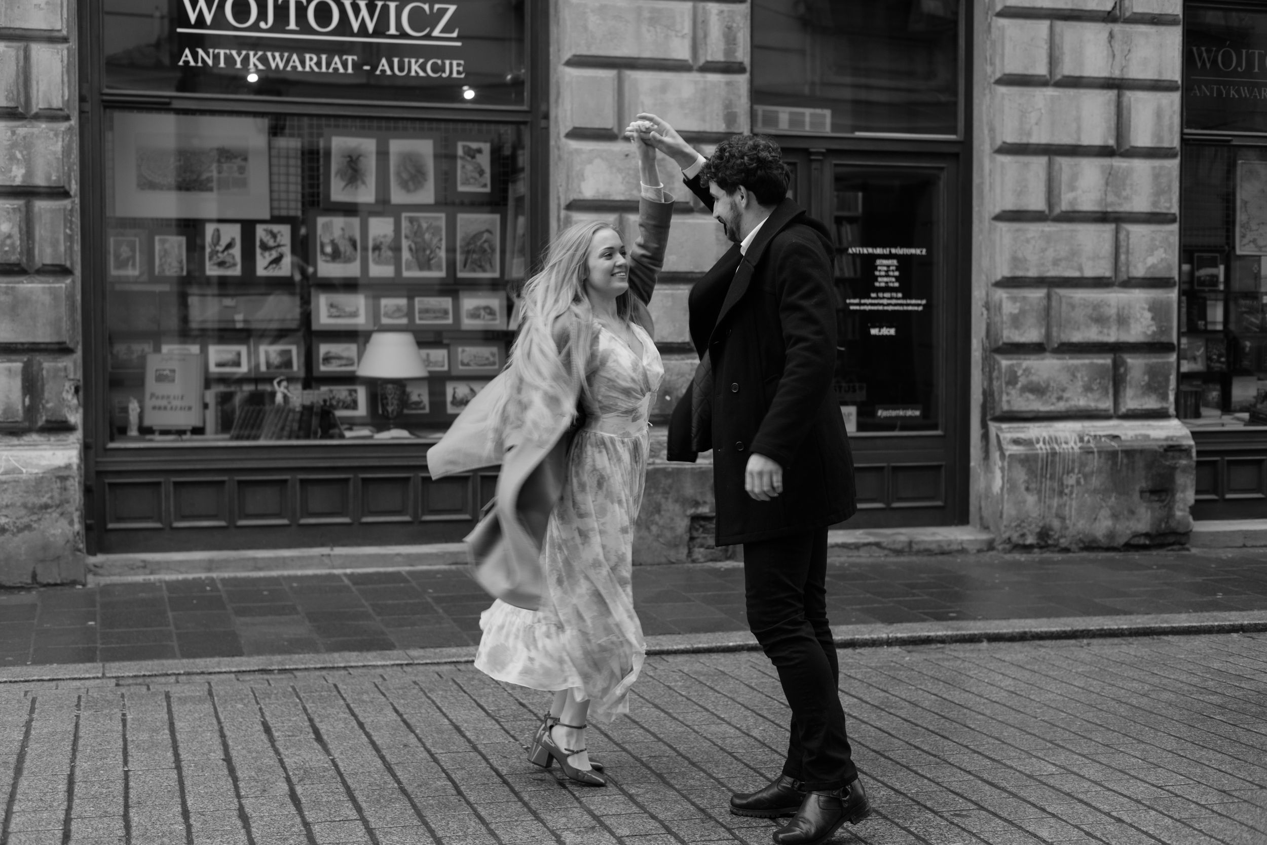 A couple in vintage wedding attire dance together on a cobblestone street in front of an antique storefront.