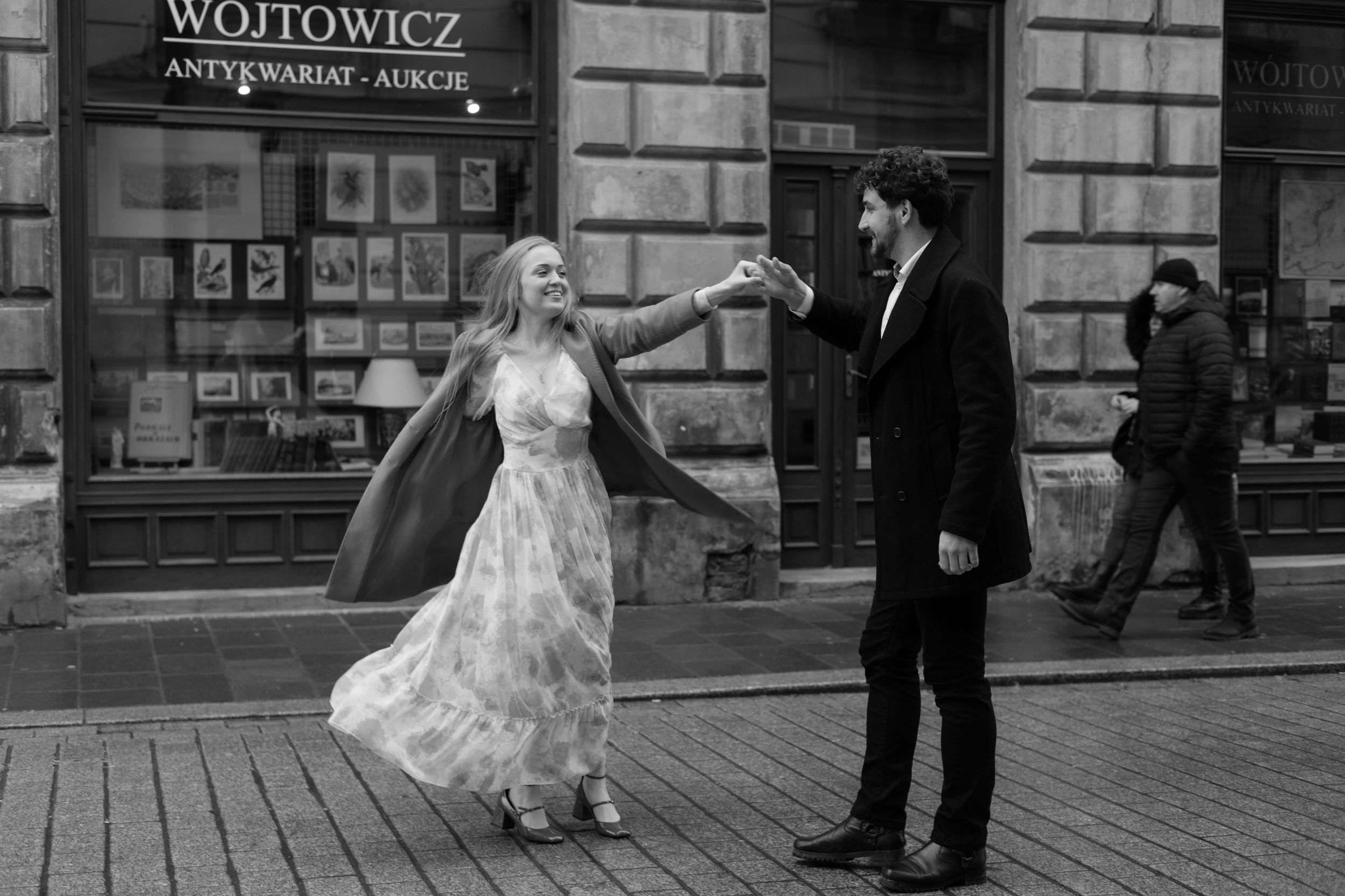 A couple in formal attire dances joyfully on a cobblestone street outside a shop in black and white photography.