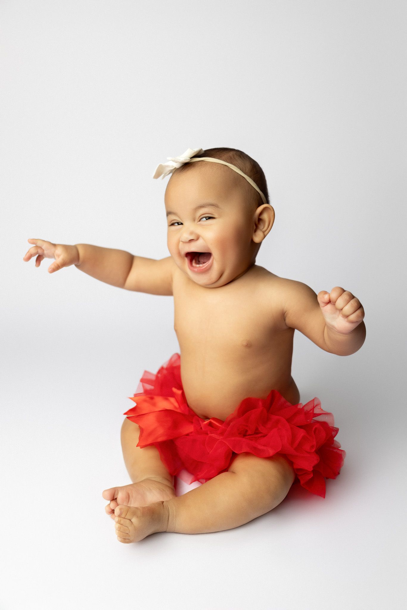Happily laughing baby wearing a red tutu skirt sits on white background while pointing their arm outward.