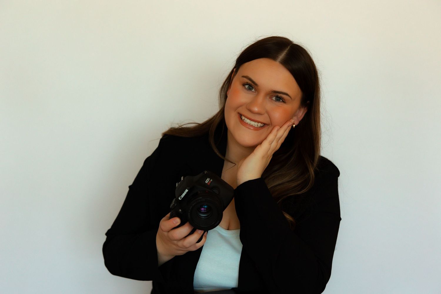 Professional photographer in black jacket holds DSLR camera while posing against white wall.