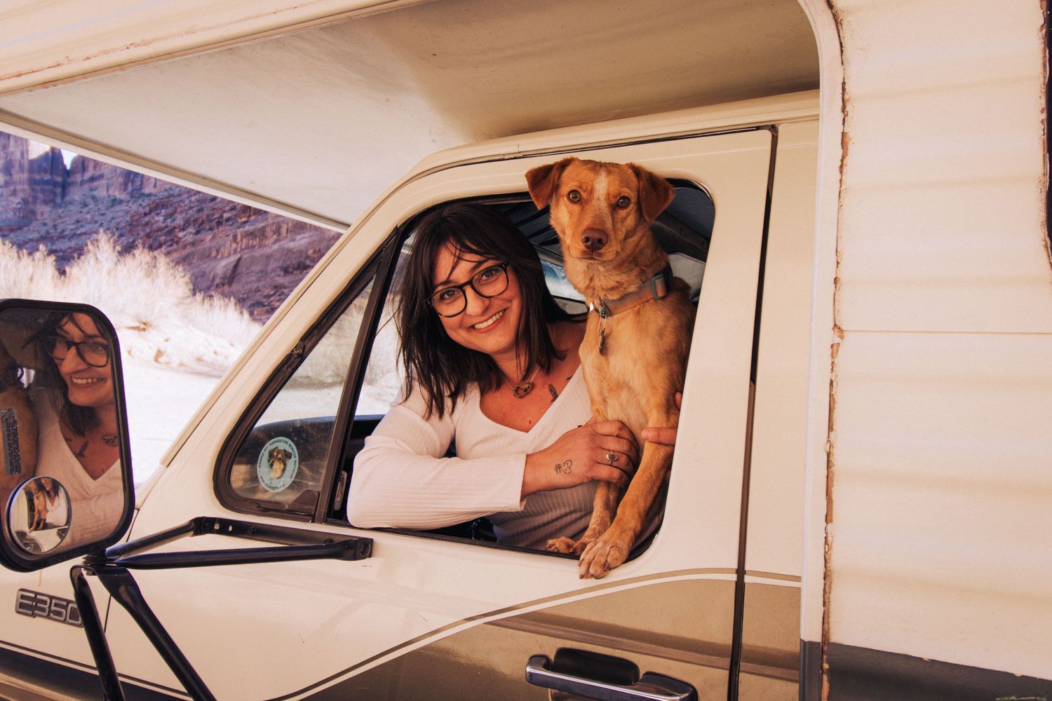 Person and dog sit together in white van window with mountains visible in background.