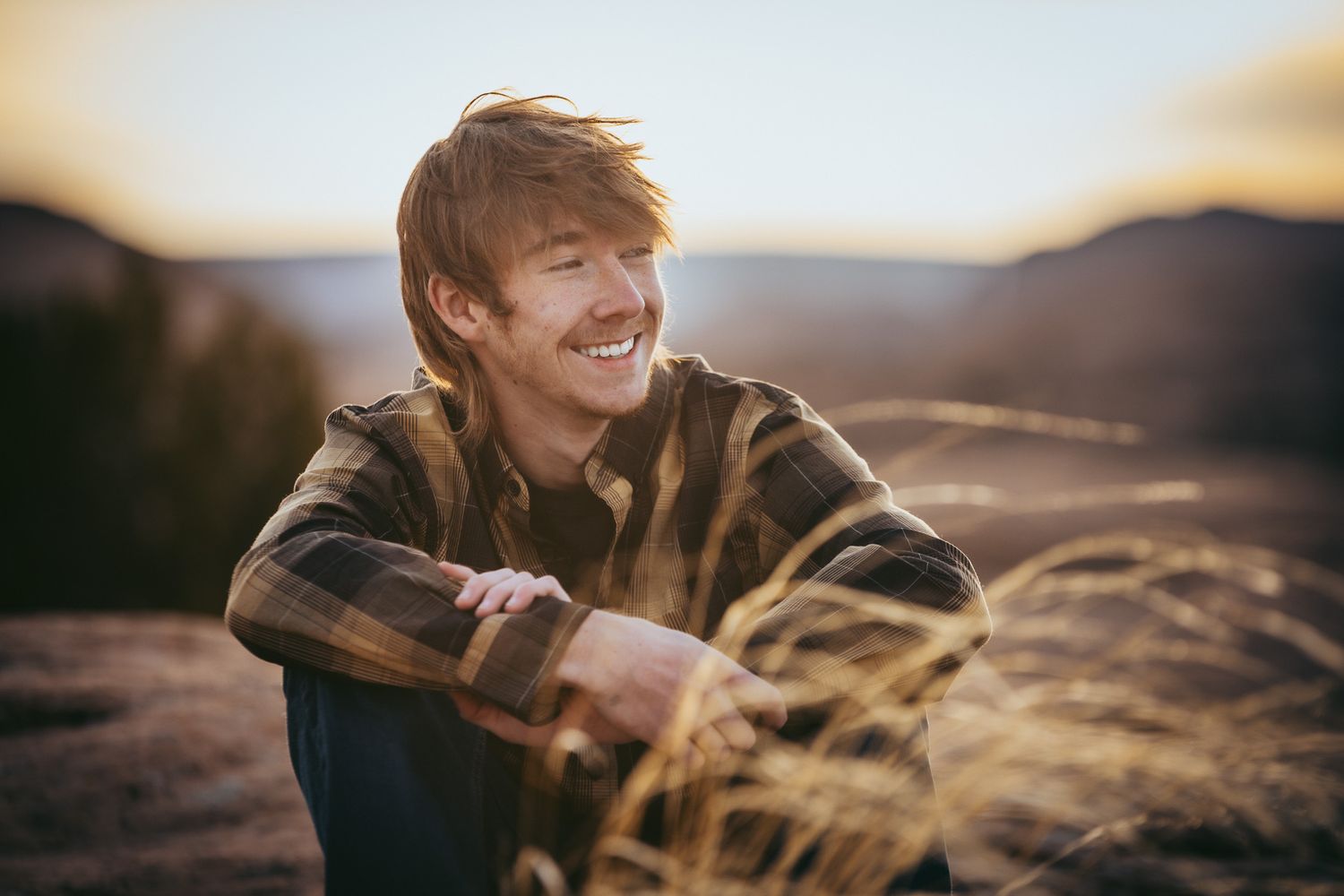 Portrait of person sitting in golden field at sunset with mountains in background.