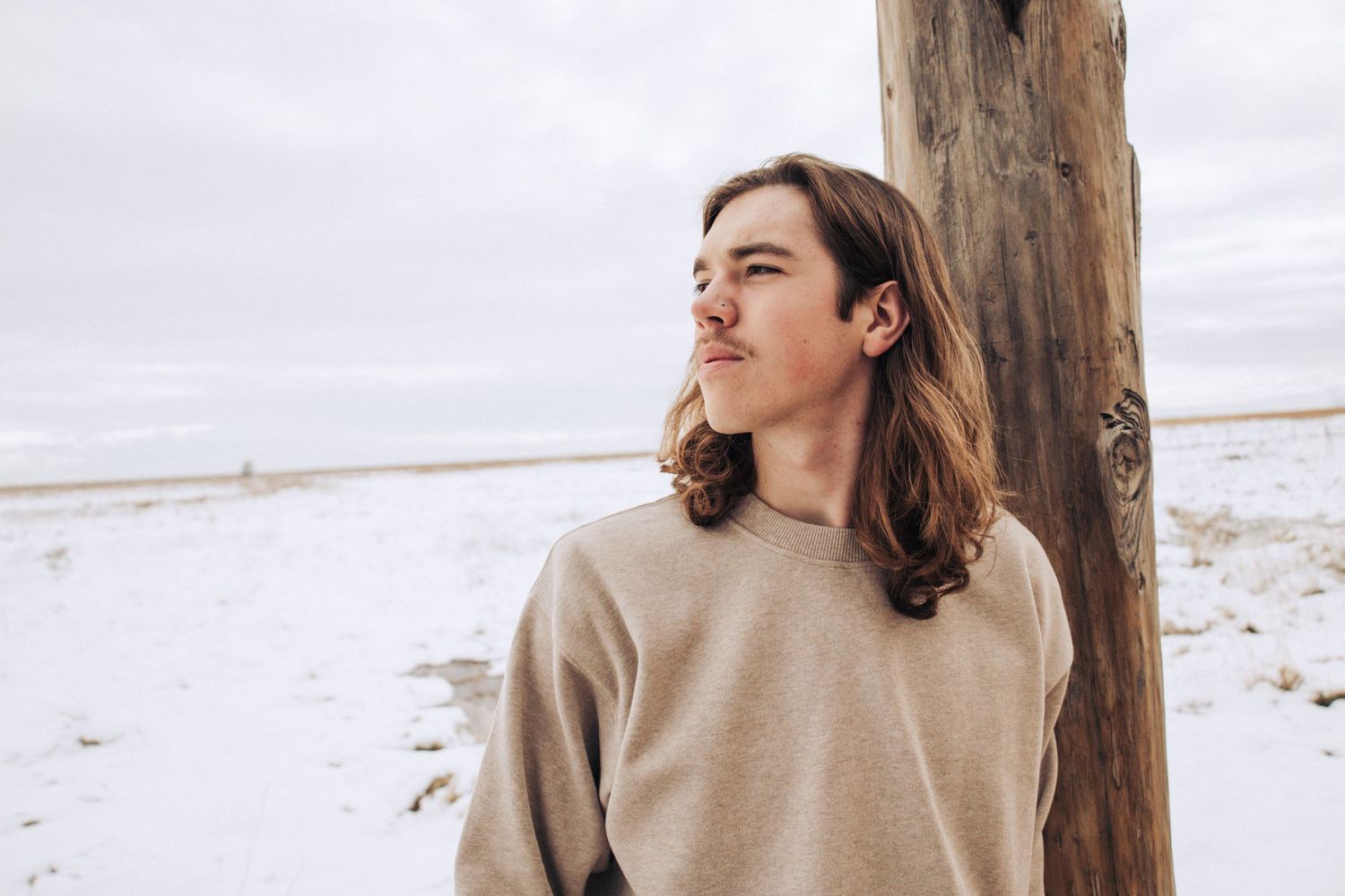 Person in beige sweater leaning against wooden post in snowy landscape.