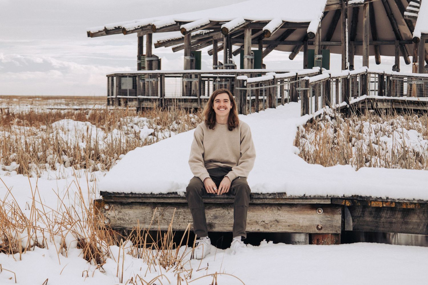 Person sitting on a snowy wooden dock near reeds and architecture in a wintry landscape.