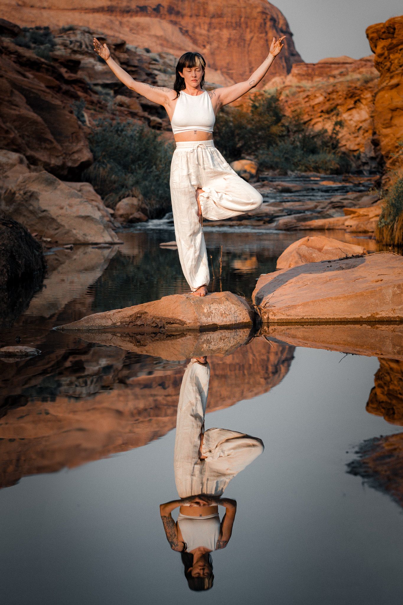 Person in white outfit balances on rock by still water creating perfect reflection in desert landscape.