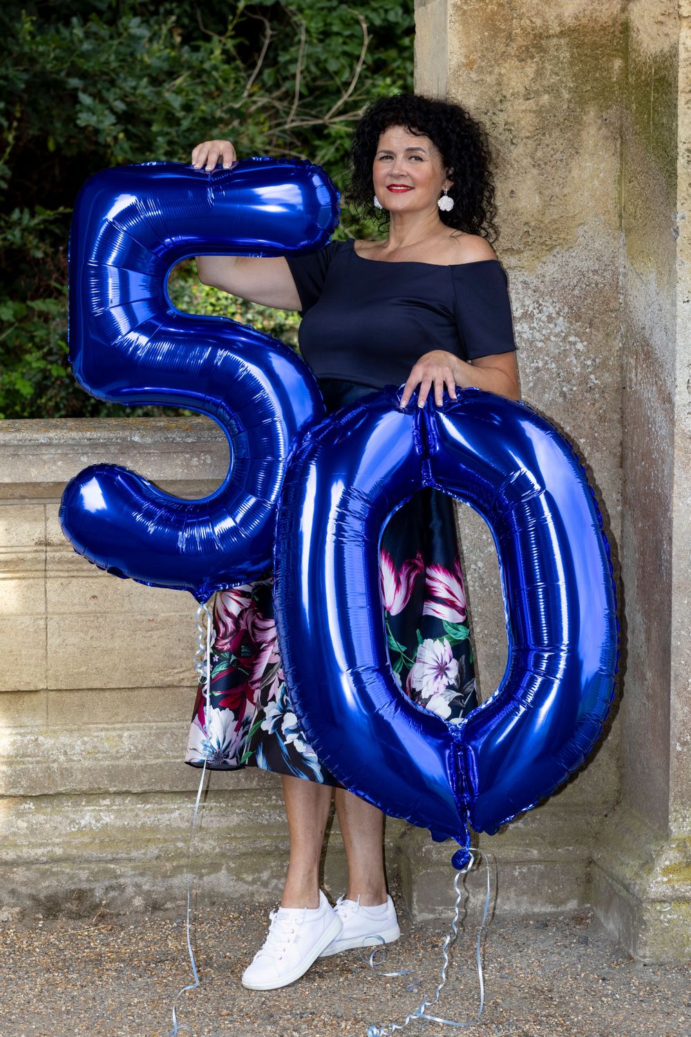 Someone wearing a navy dress poses with large blue number 50 balloons against a stone wall.