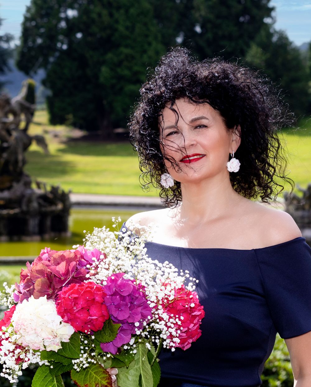 A person in a navy off-shoulder dress holds a vibrant bouquet of pink flowers in a garden setting.