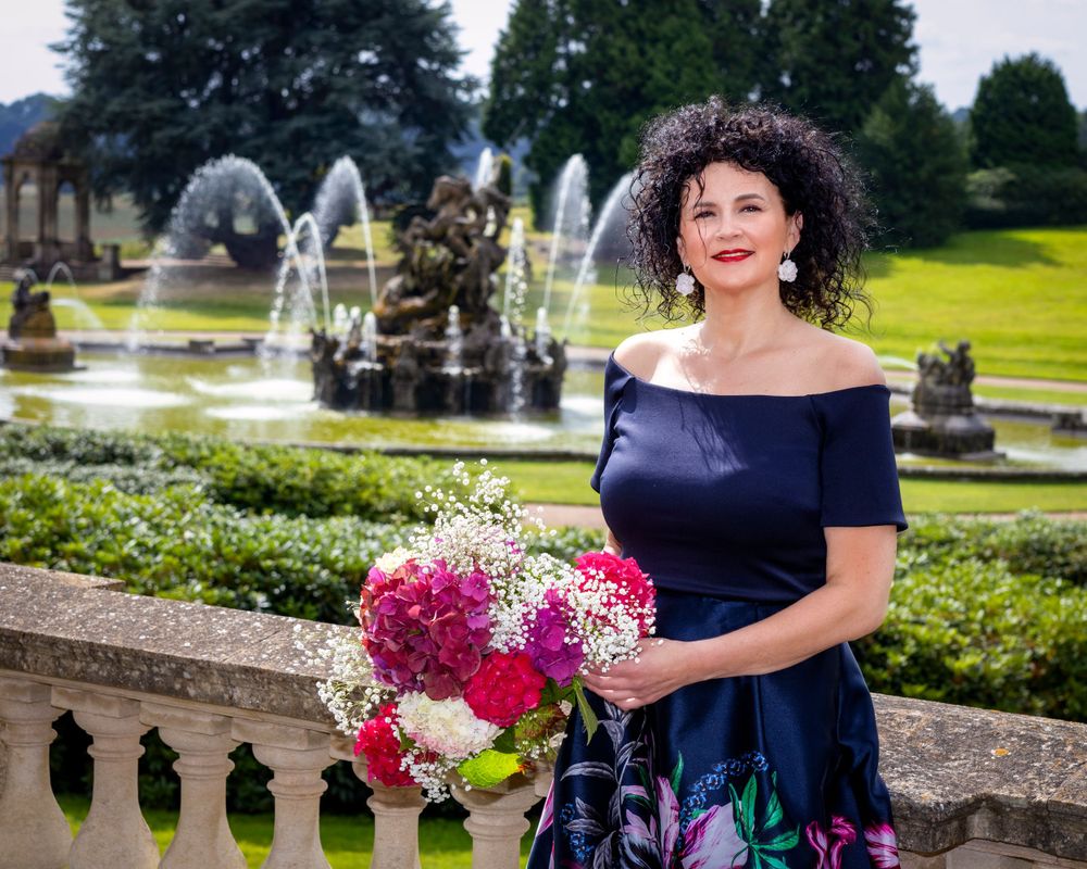Person in navy dress holds flowers while posing by ornate fountain in formal garden setting.
