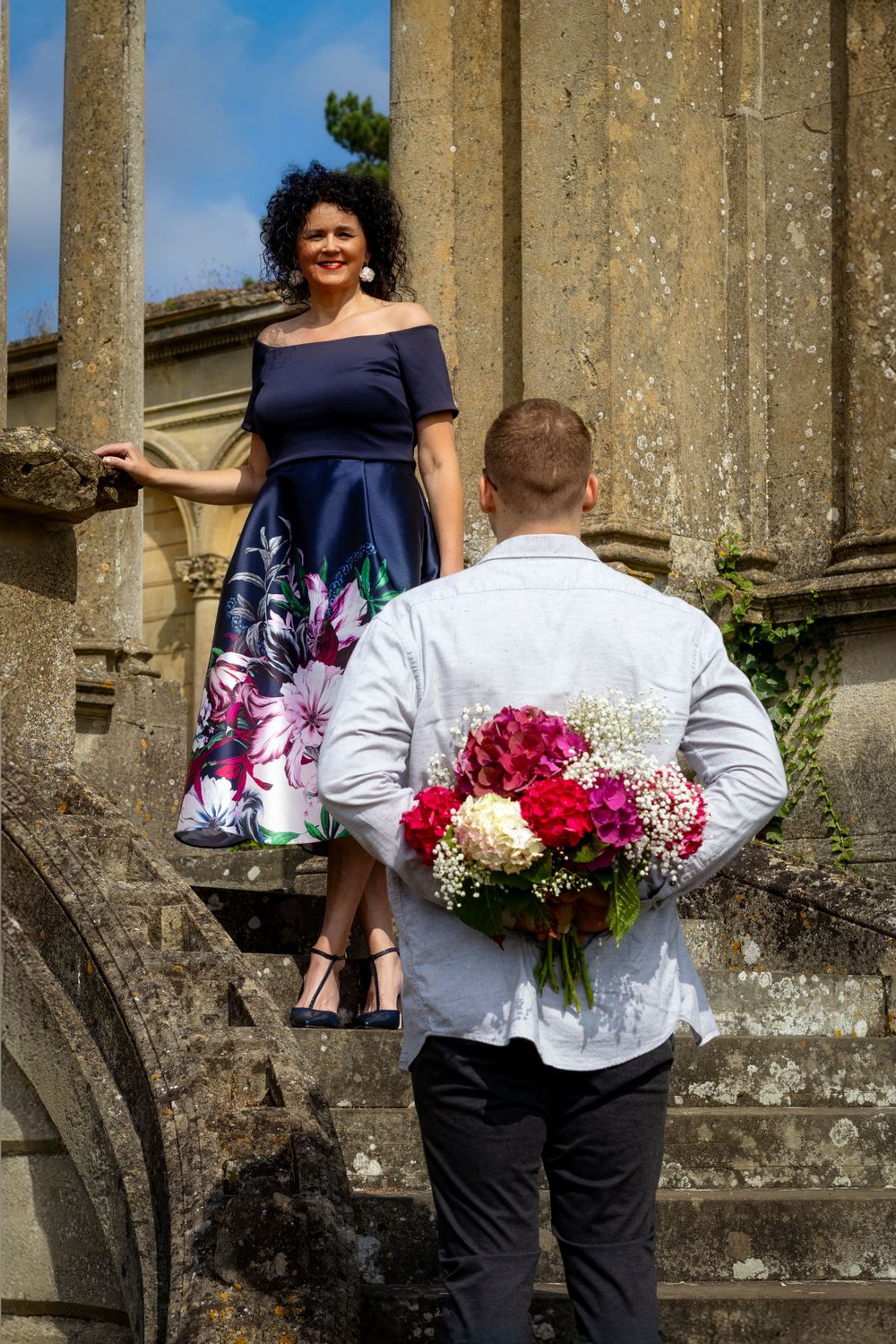 Person on stone steps while someone in white shirt holds flowers behind their back.