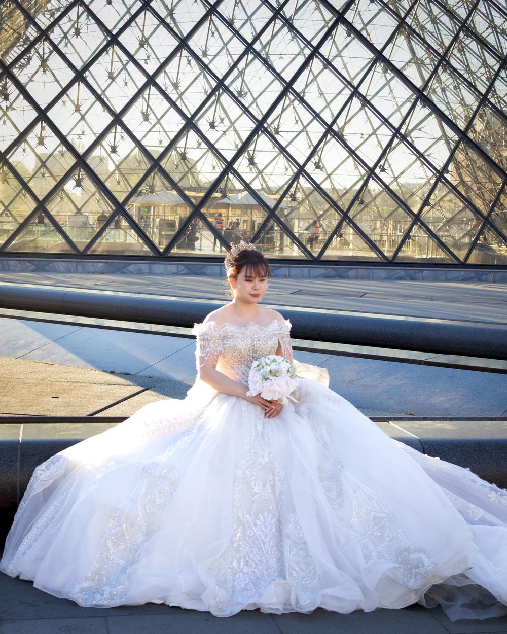 An elegant bride in a luxurious white ballgown poses at the iconic glass pyramid entrance of the Louvre Museum in Paris.