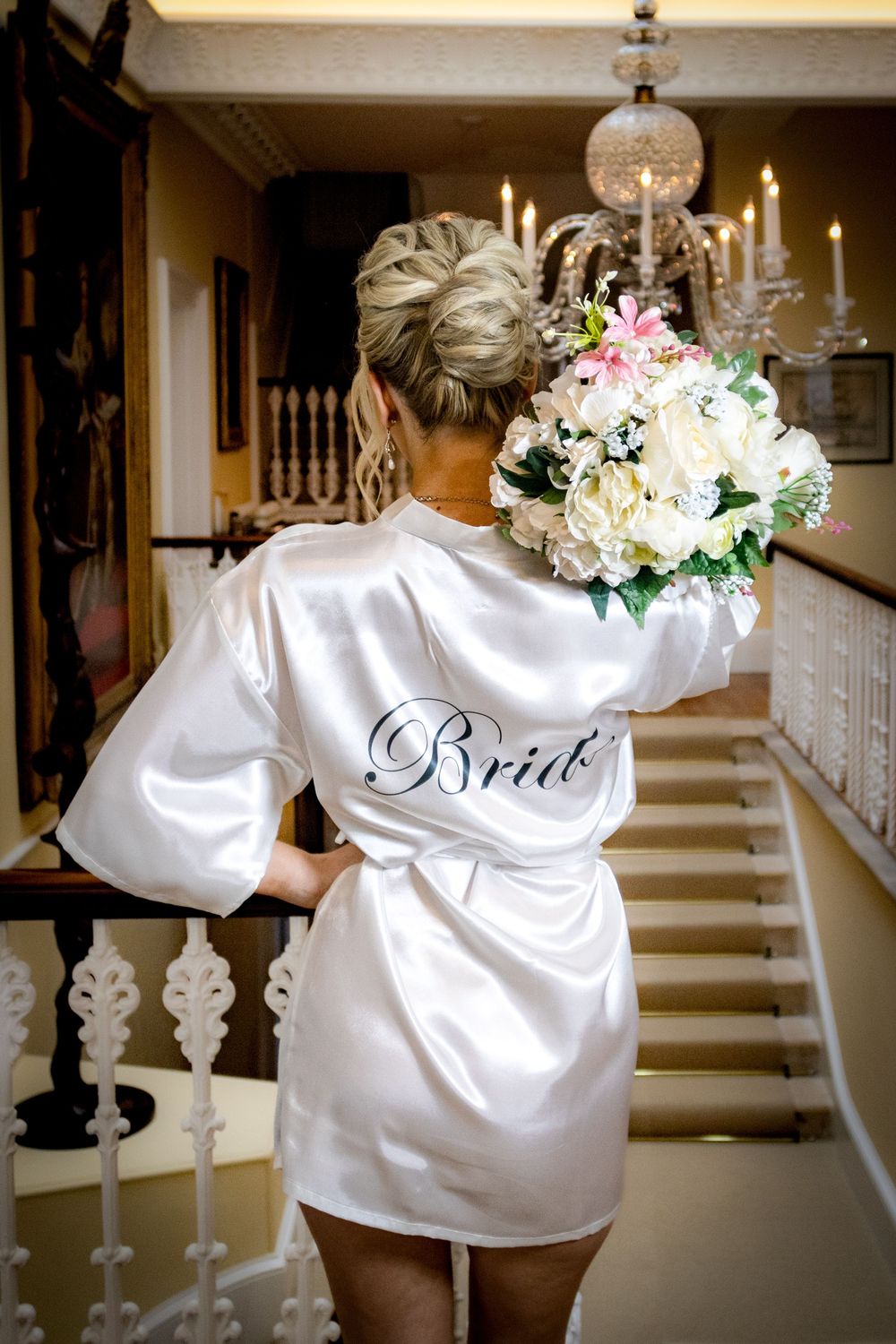 A person wearing a white silk robe with 'Bride' embroidered on the back holds a bouquet by an elegant staircase.