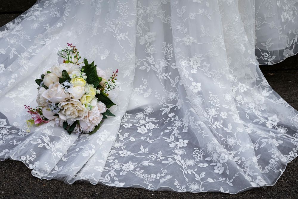 A delicate bridal bouquet of white flowers rests on flowing lace wedding dress fabric with floral embroidery details.