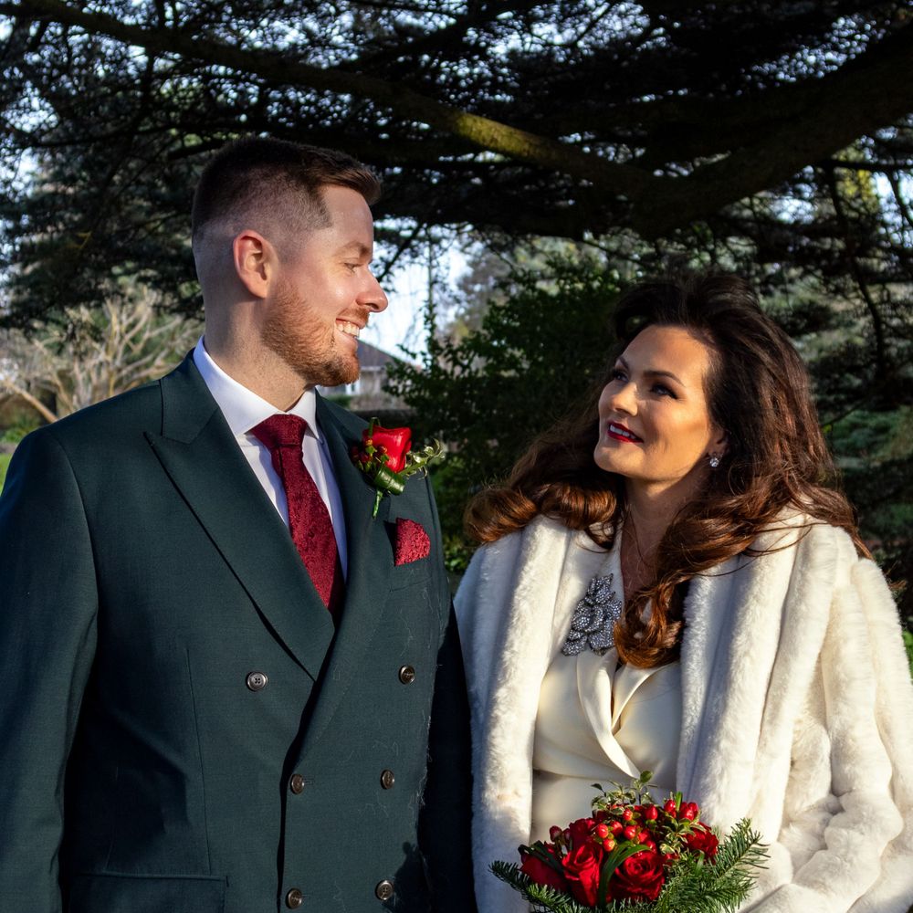 A couple in wedding attire share a moment outdoors with evergreen trees in background.