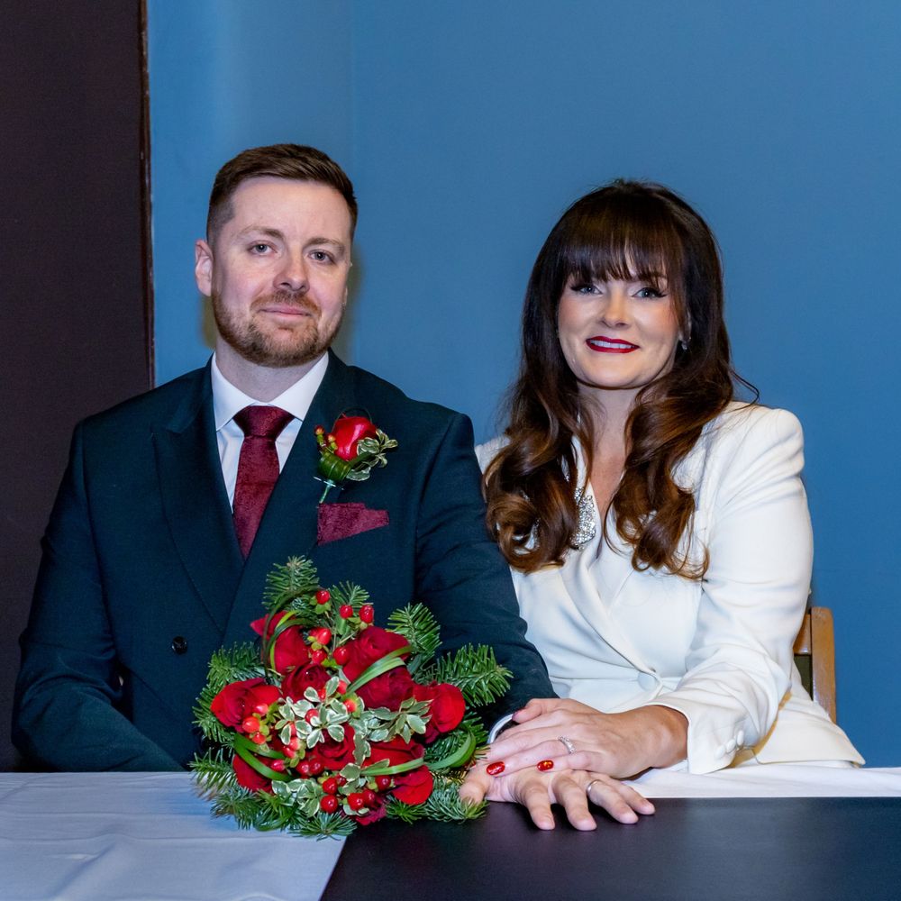 A couple in formal attire sits together holding a red rose bouquet.