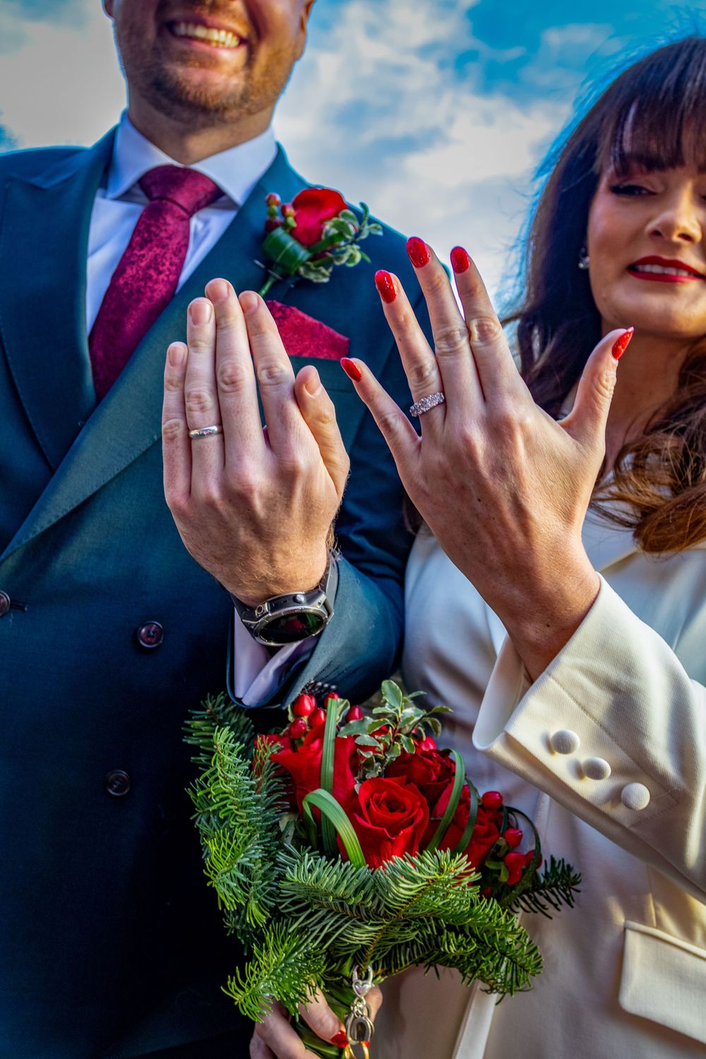 Newlyweds display their wedding rings while holding a festive red rose bouquet.