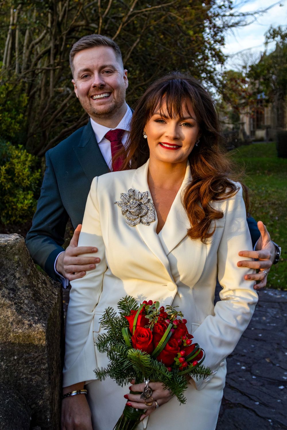 A couple in formal winter wedding attire poses with a red rose bouquet.