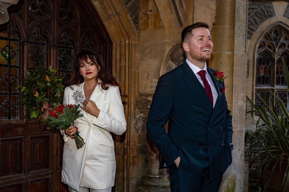 A couple in formal attire poses playfully in front of gothic architecture.