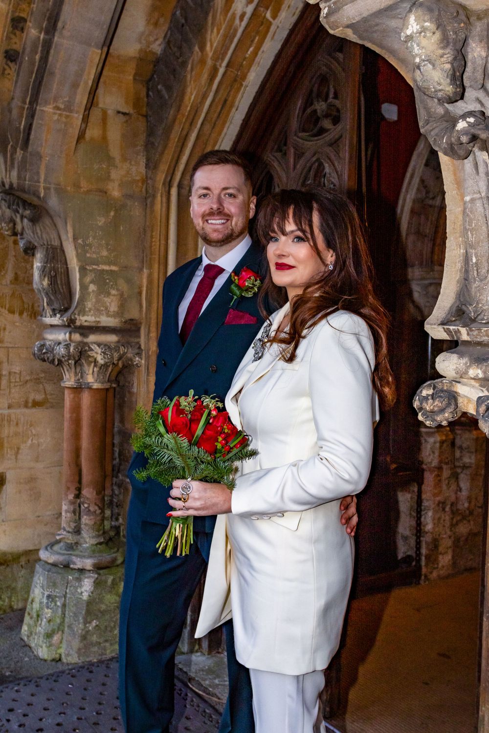 A couple poses in formal attire under ornate stone arches with a red rose bouquet.